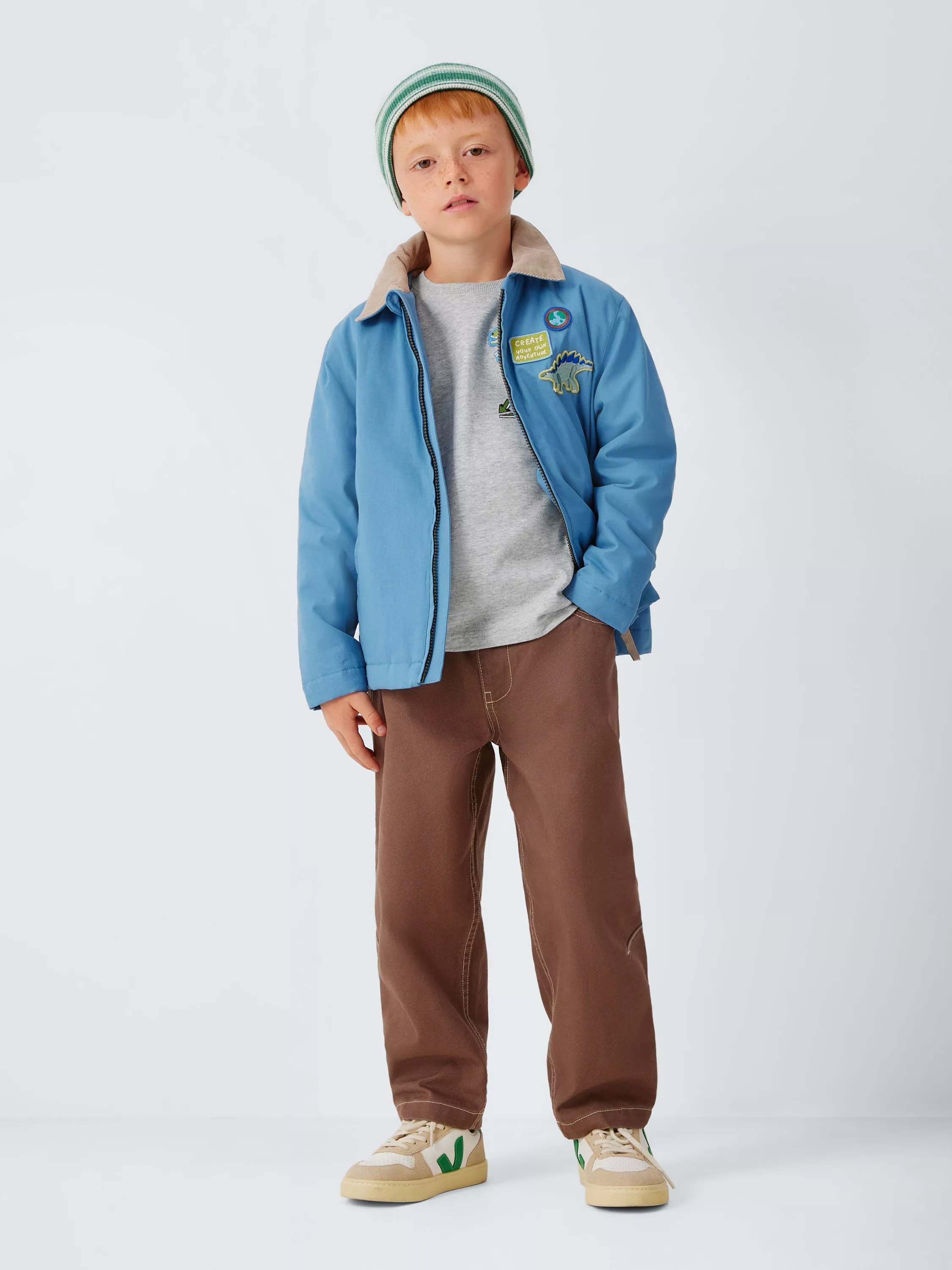 Boy in blue jacket with dinosaur patch, grey T-shirt, brown trousers and beanie, studio fashion photo