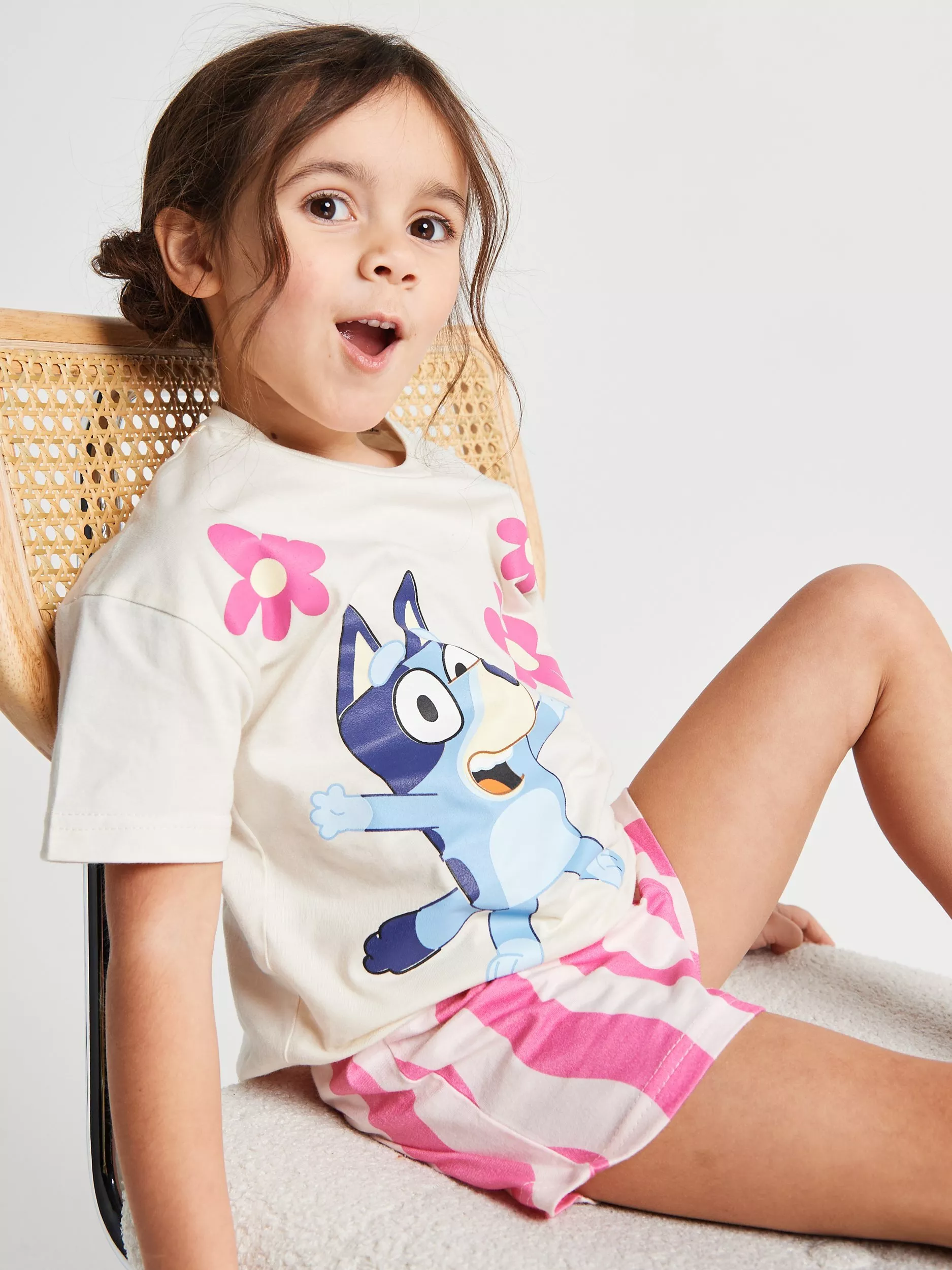 Girl wearing Bluey graphic T-shirt and pink shorts, sitting on a rattan chair in a studio portrait setting