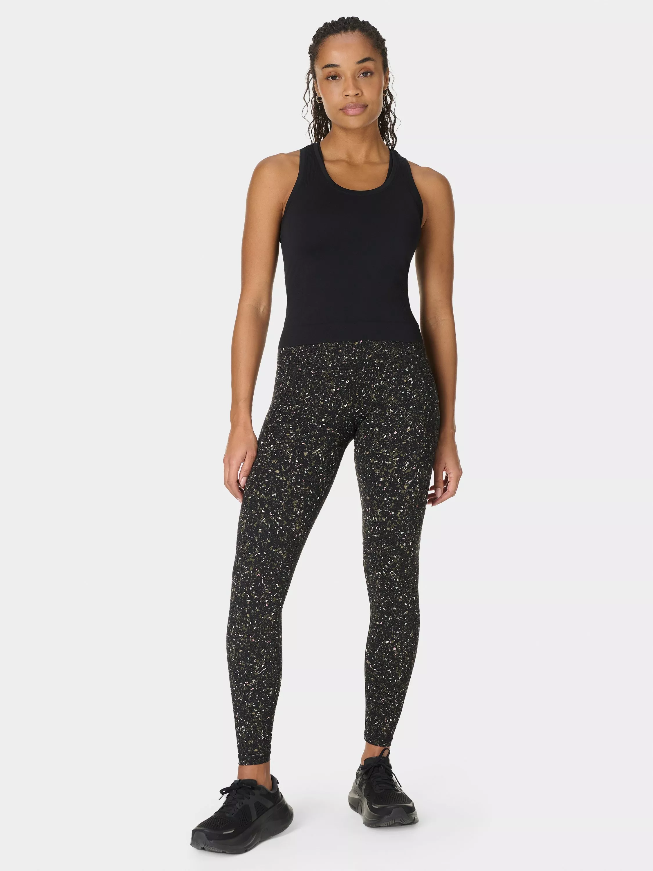 Full-length studio photo of woman wearing black racerback bodysuit and speckled high-waist leggings with trainers