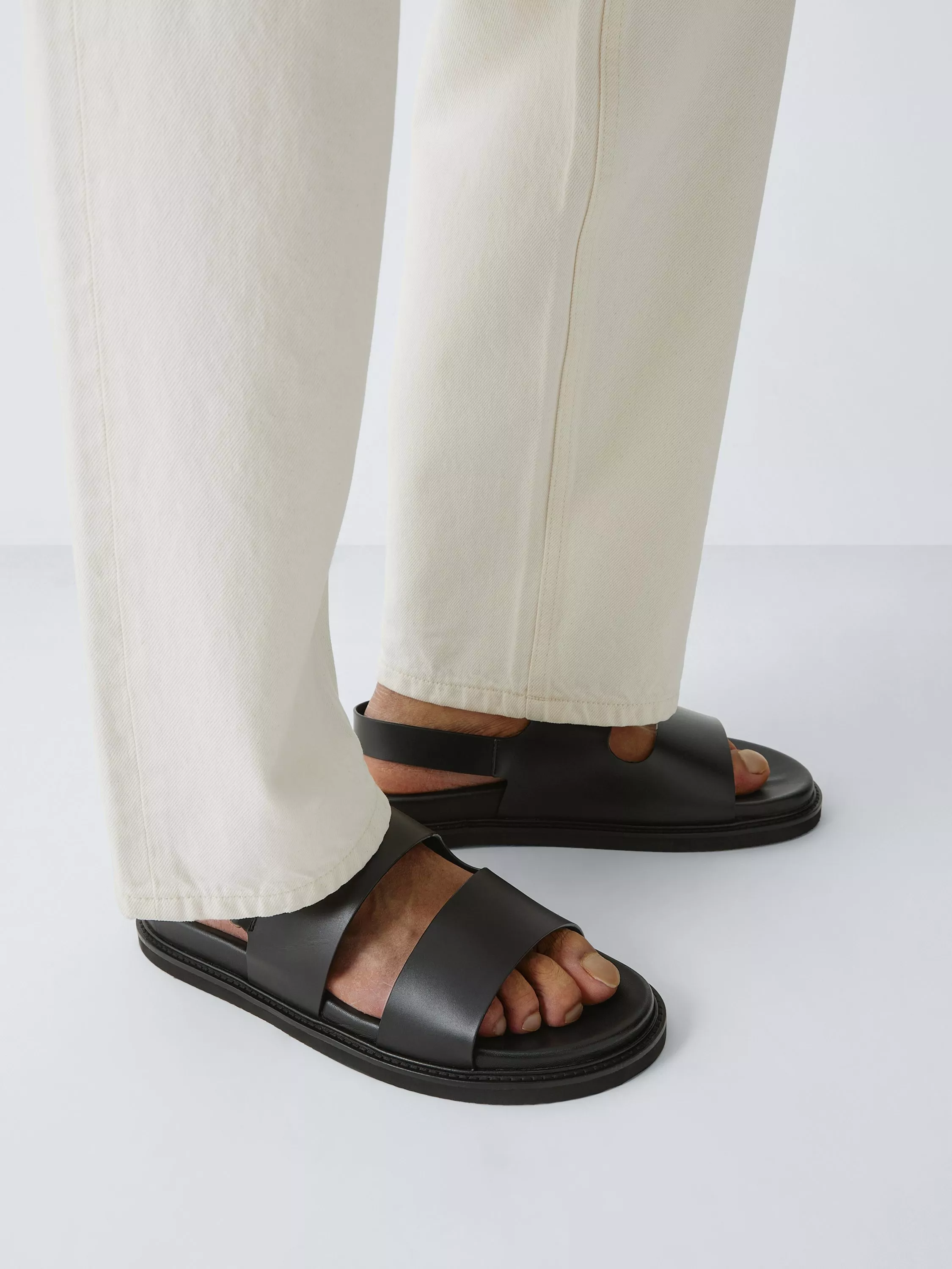 Close-up of black leather slingback sandals with wide straps, worn with cream trousers on a studio backdrop