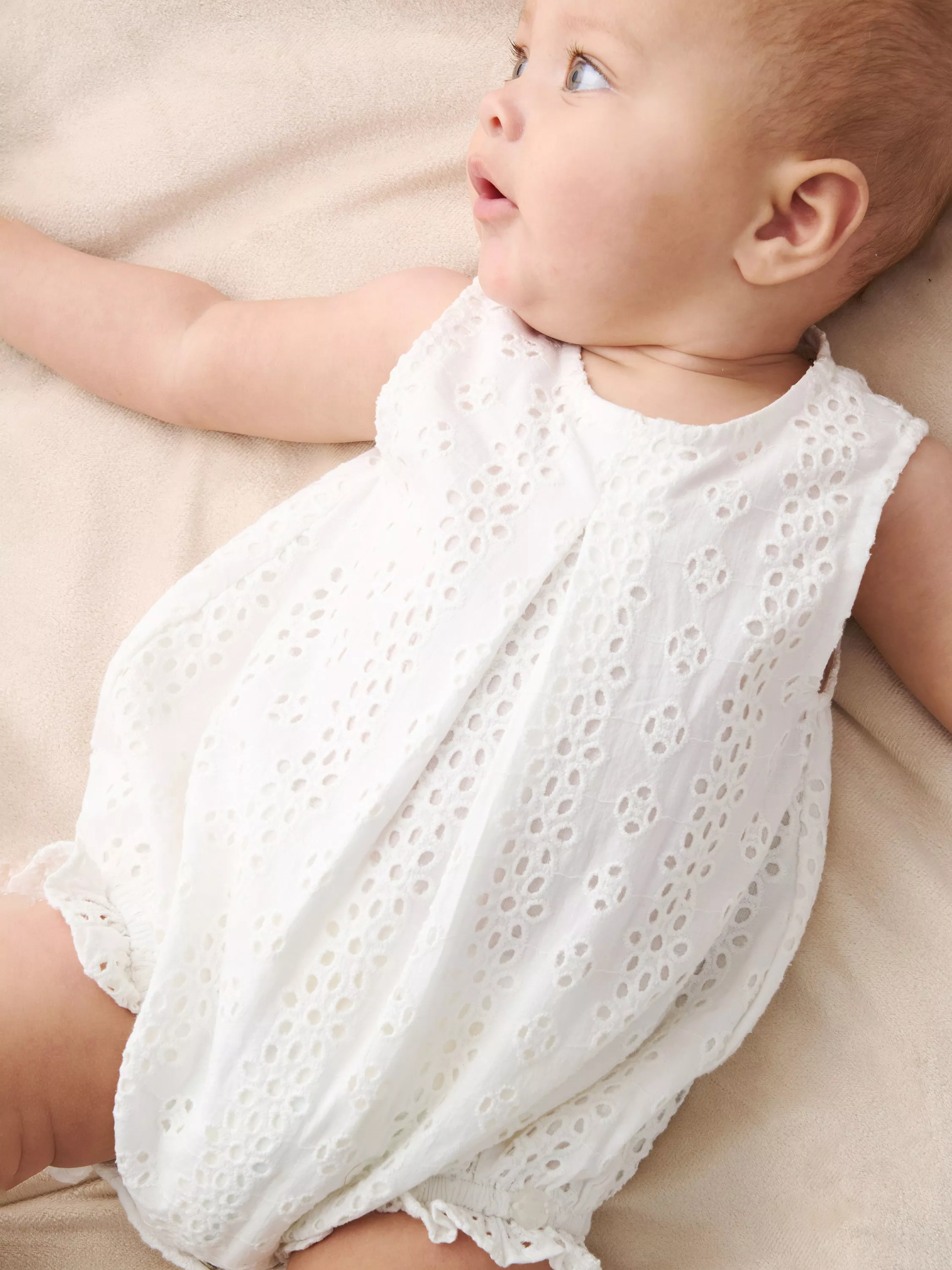 Baby in white broderie anglaise romper lying on beige blanket, soft natural light newborn portrait