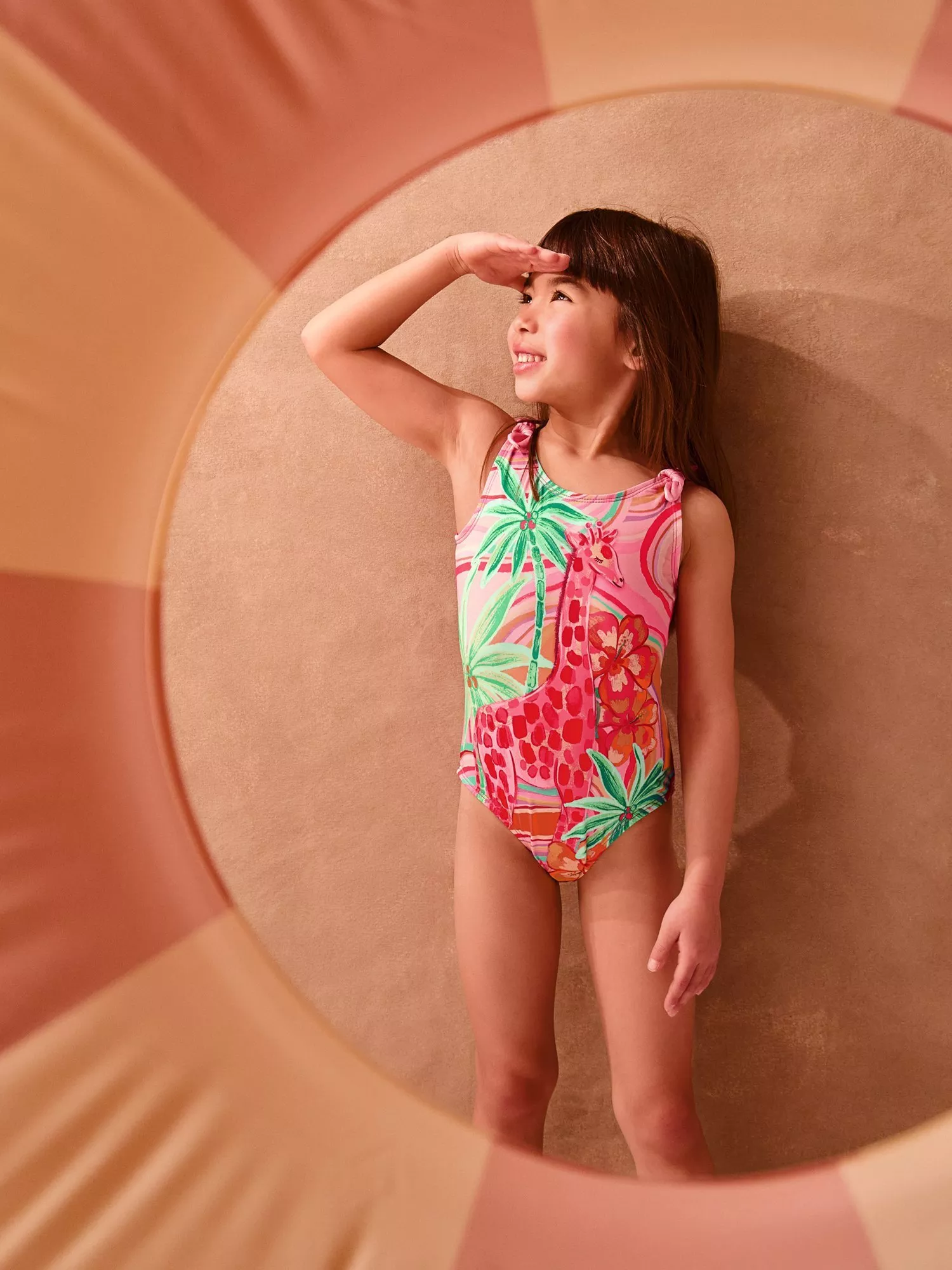 Smiling girl in colourful giraffe and palm print swimsuit, shading eyes, standing in striped beach cabana doorway.