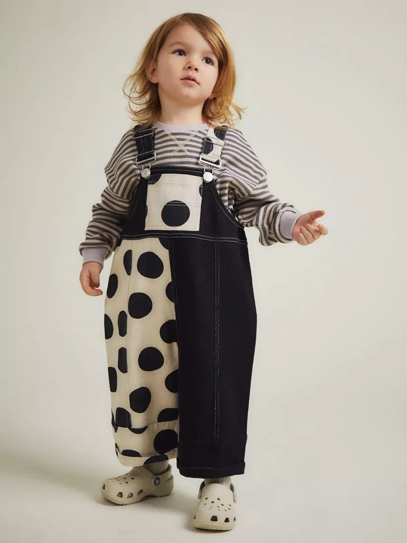 Toddler in striped top and black-and-cream polka dot dungarees, standing in studio on neutral backdrop