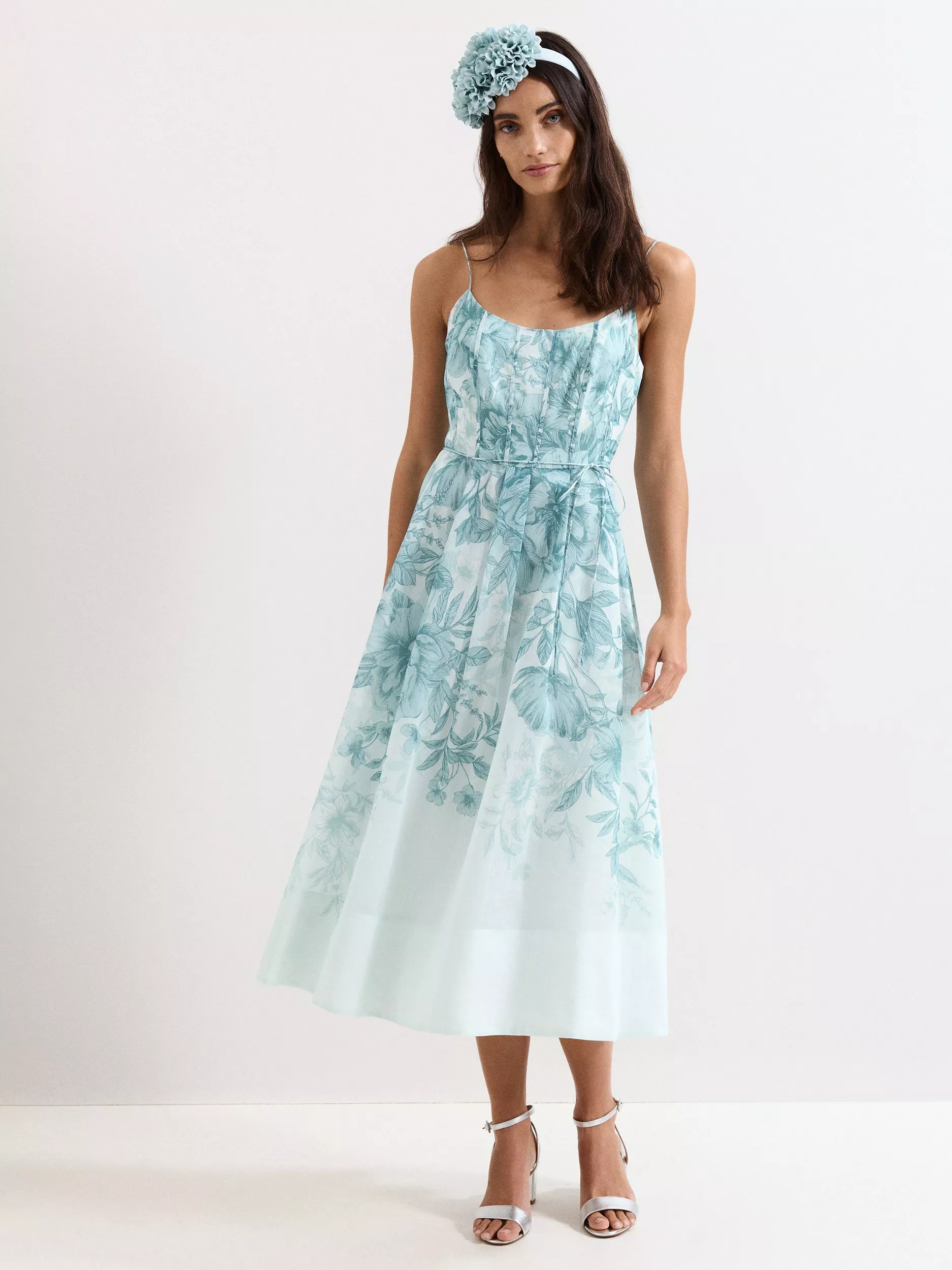 Woman in navy floral midi dress with fascinator hat, white heels and clutch, elegant wedding guest outfit.