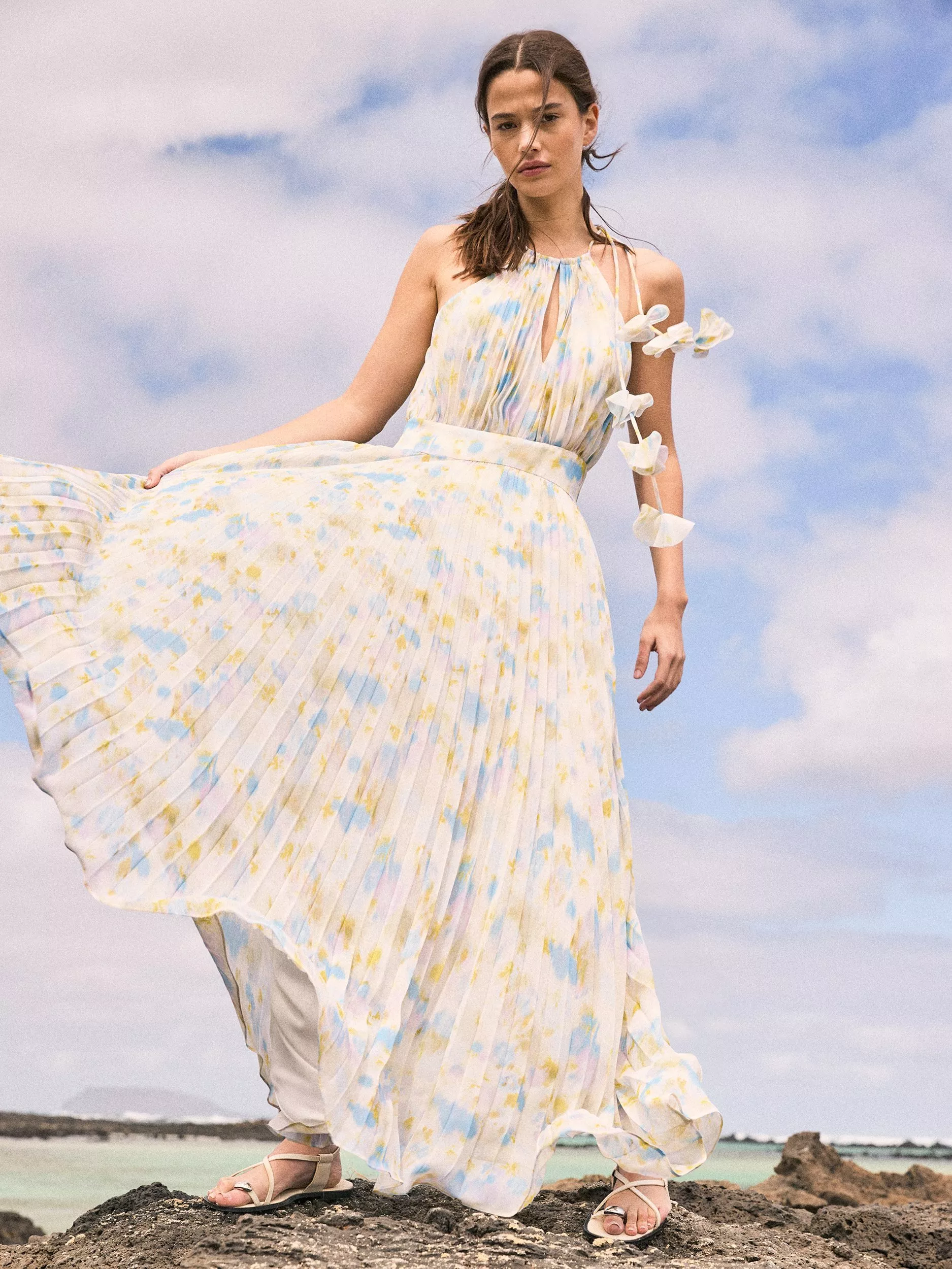 Woman modelling a flowing pastel floral maxi dress on coastal rocks, blue sky backdrop, summer fashion style