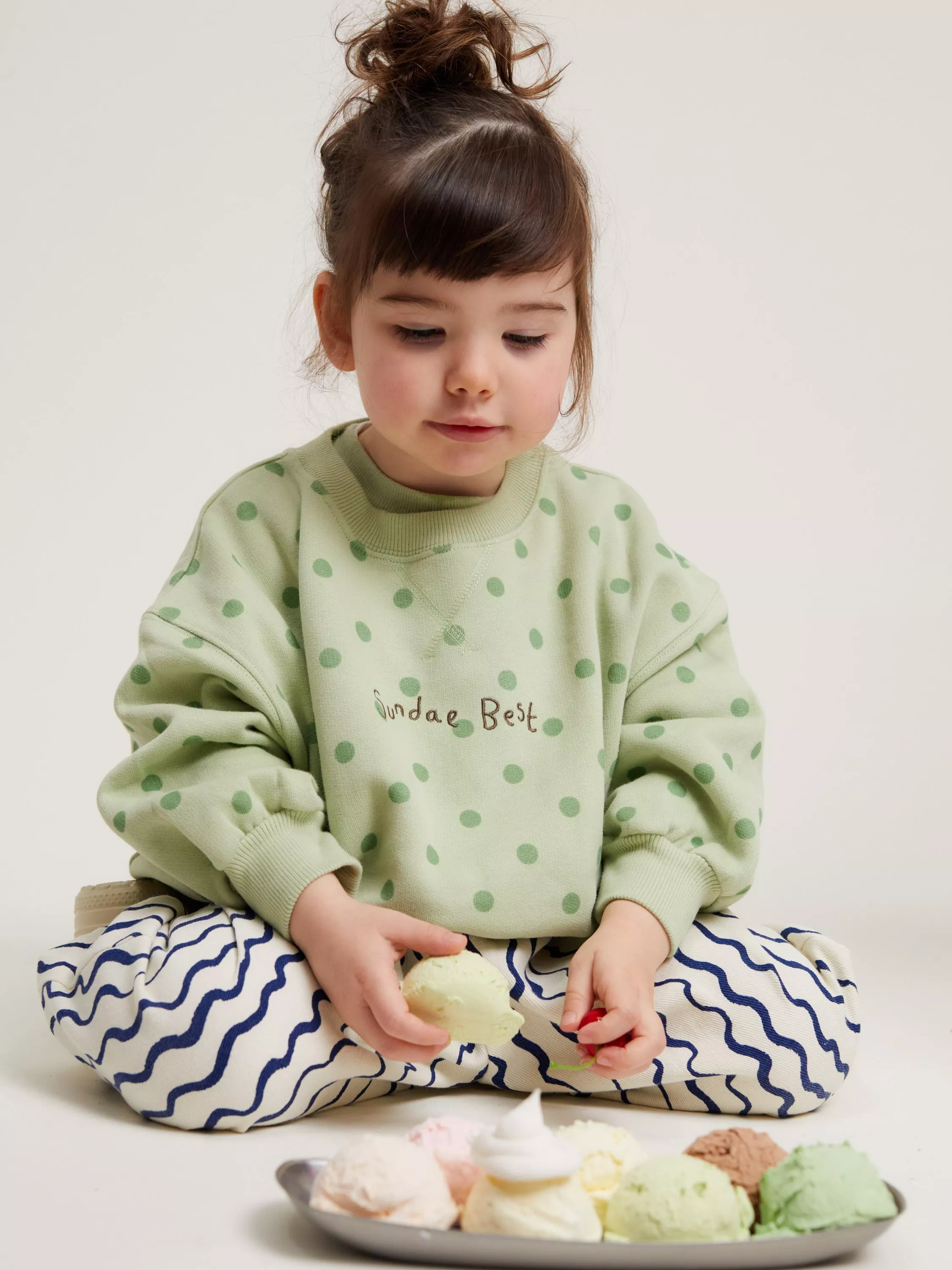 Toddler in green polka dot sweatshirt and patterned trousers sitting with colourful ice cream scoops on plate