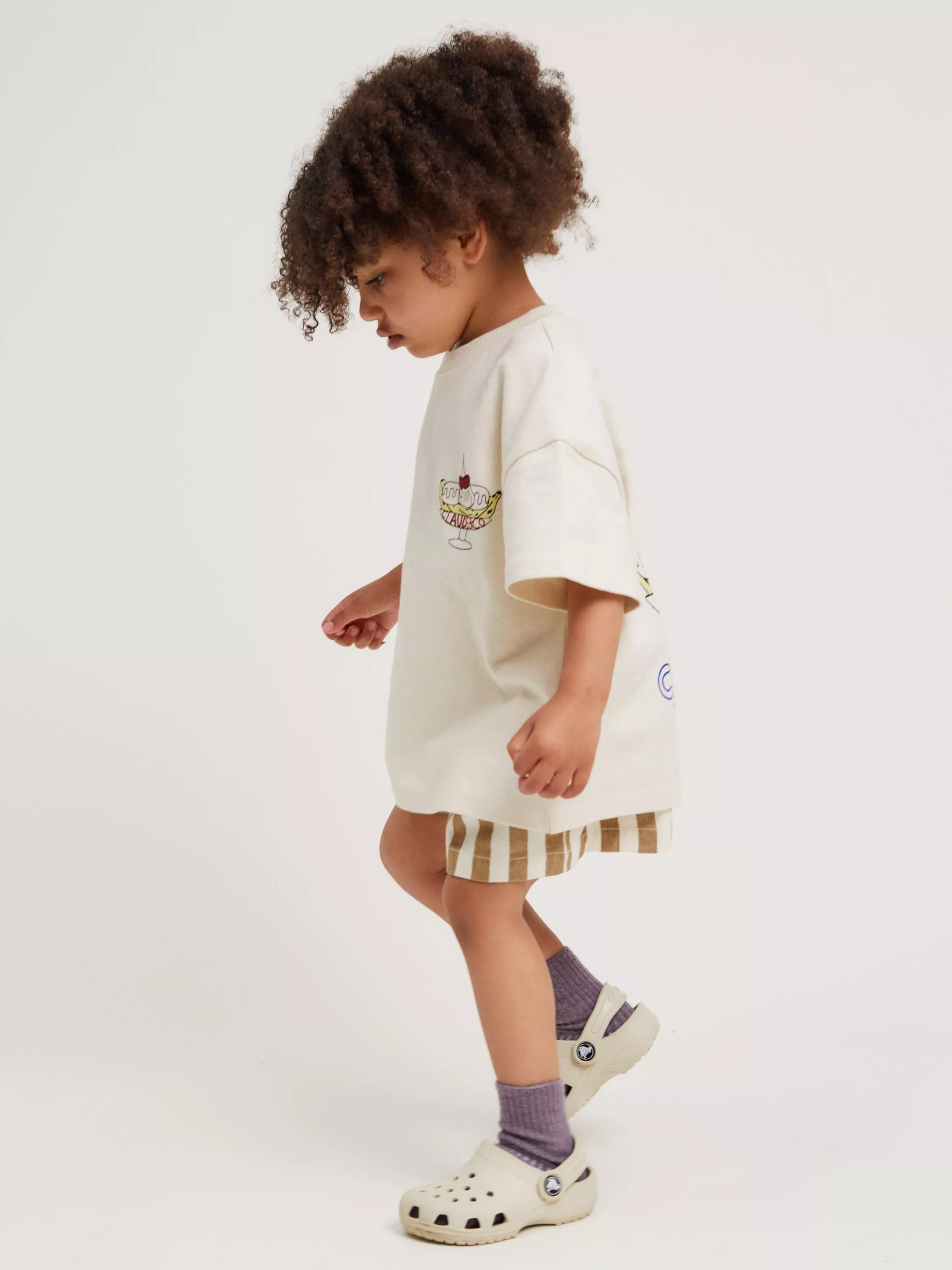 Toddler in cream graphic T-shirt and striped shorts, wearing purple socks and white clogs, on white backdrop
