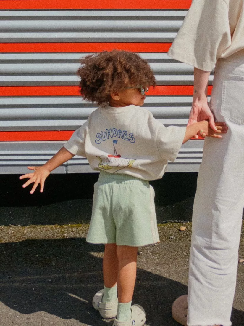 Toddler holding an adult’s hand outdoors, wearing a printed T-shirt, shorts and sunglasses by a striped wall
