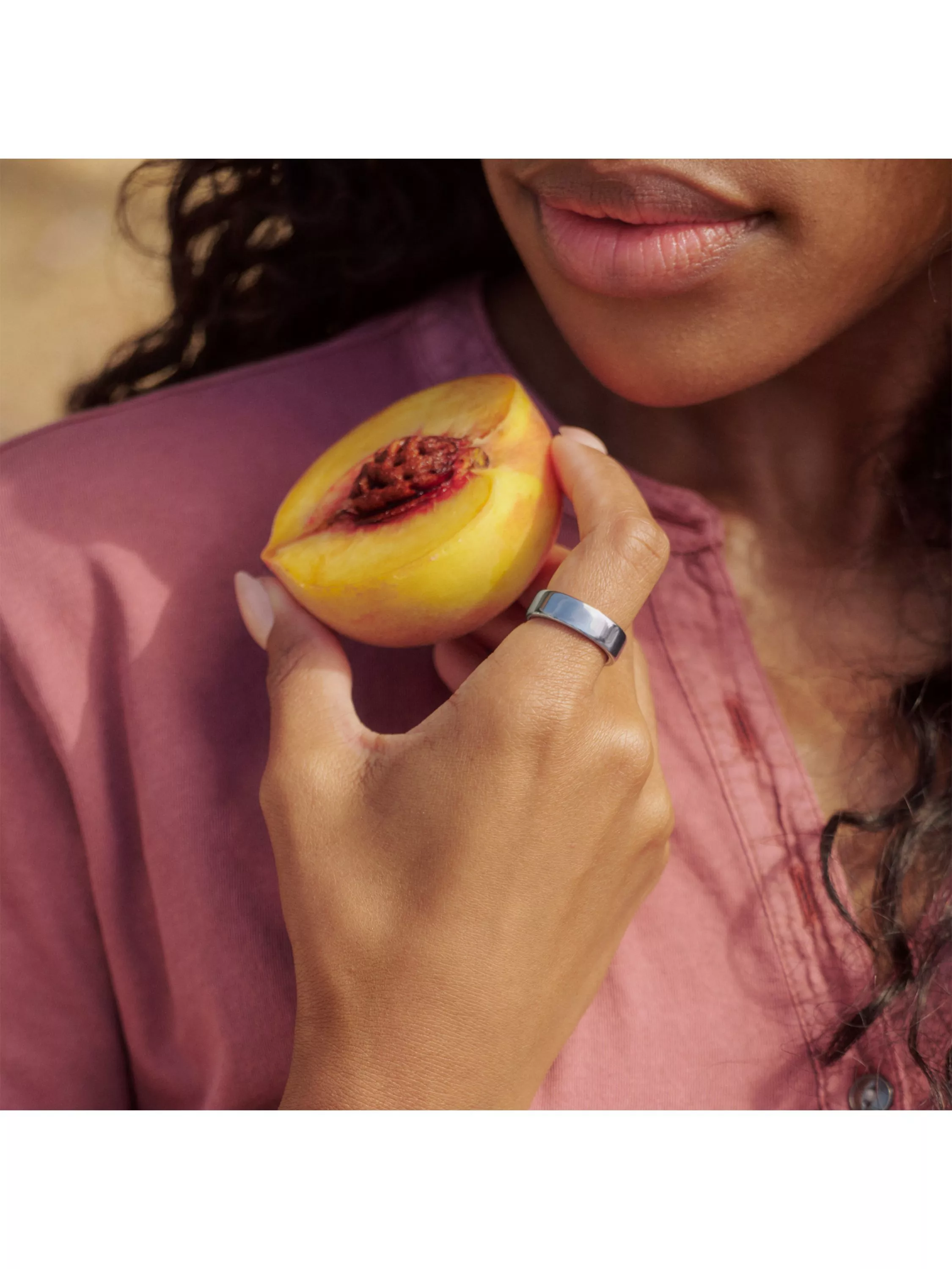 Close-up of woman holding a ripe peach near her lips, wearing a silver ring and pink blouse outdoors