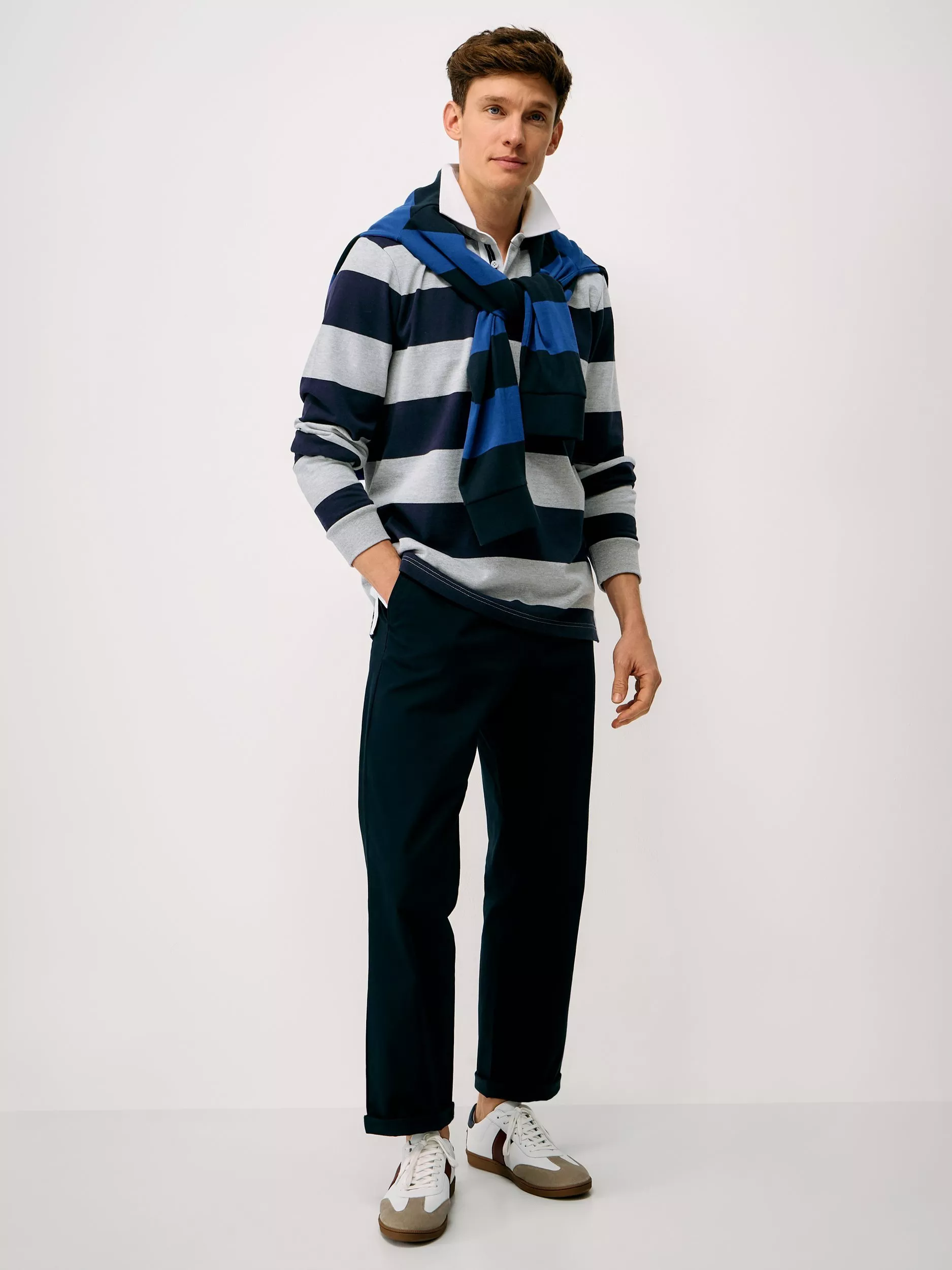 Men’s navy striped rugby shirt with blue jumper over shoulders, smart trousers and white trainers, studio shot