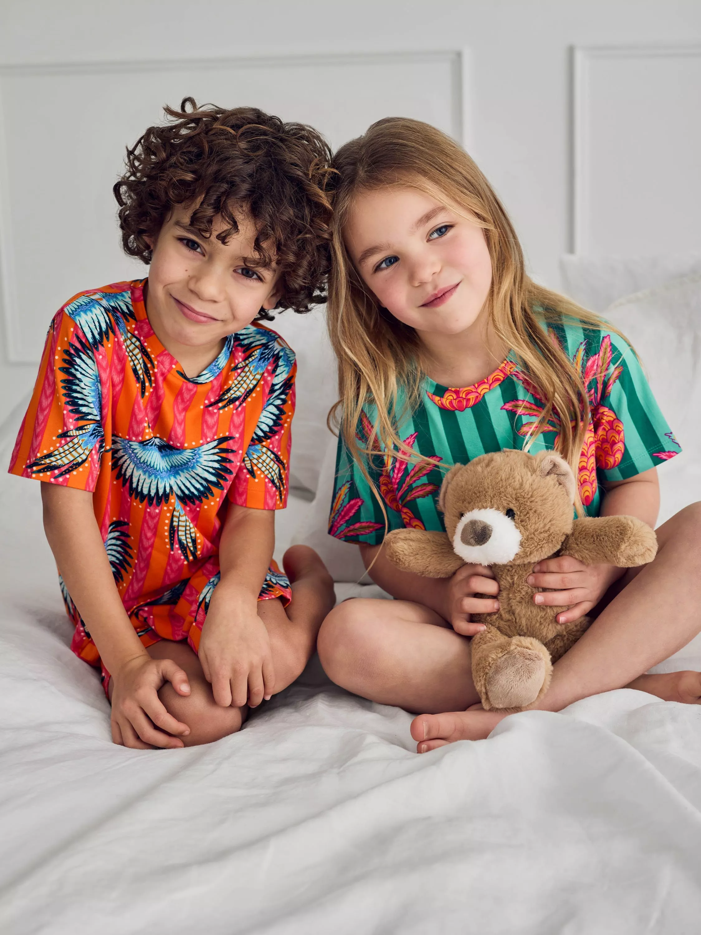 Two children in colourful pyjamas sitting on a white bed, girl holding a teddy bear, smiling indoors