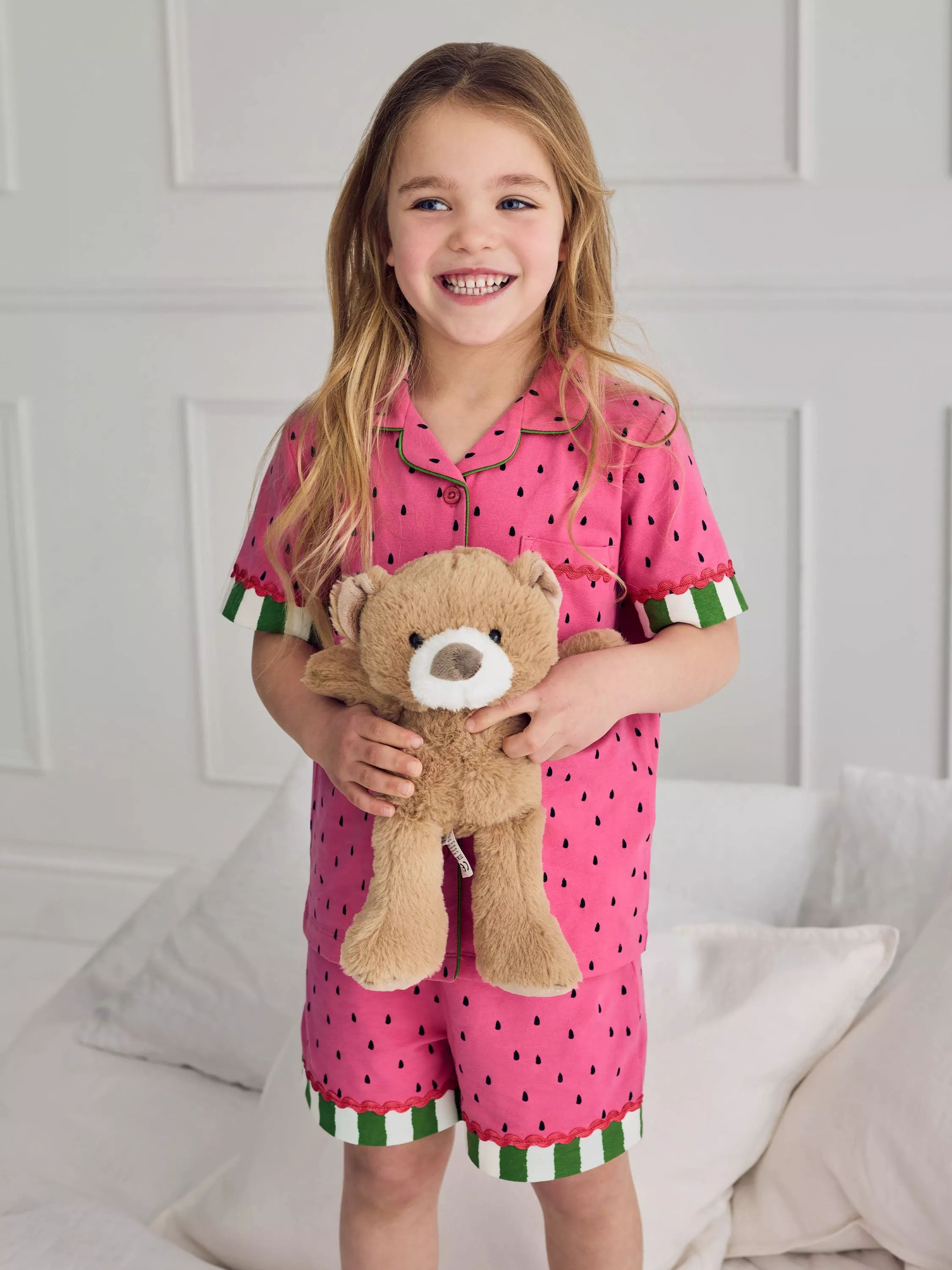 Smiling girl in pink watermelon-print short pyjamas holding a teddy bear, cosy kids bedtime loungewear indoors