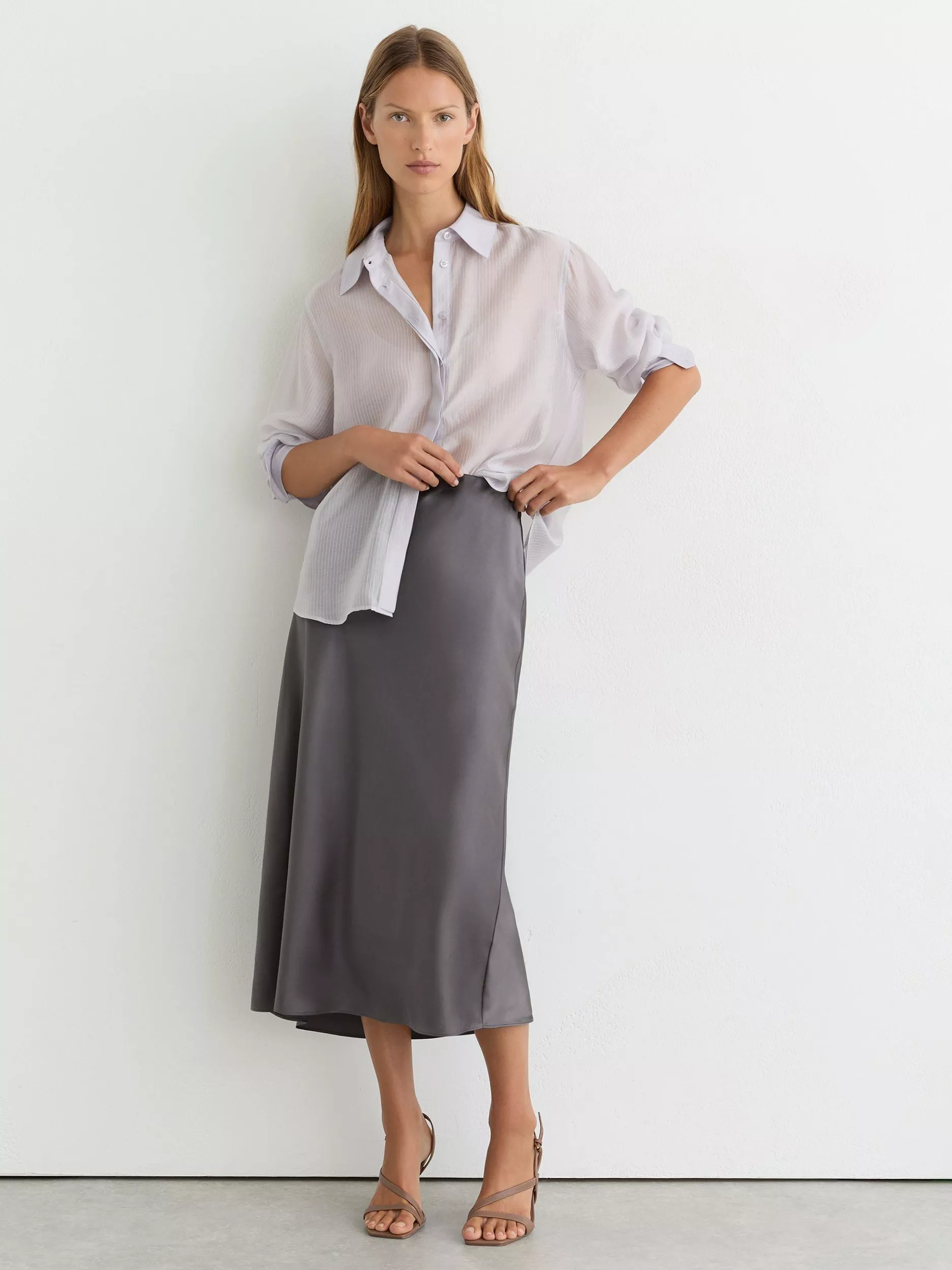 Model wearing a light lilac oversized button-up shirt with a charcoal satin midi skirt and strappy heels
