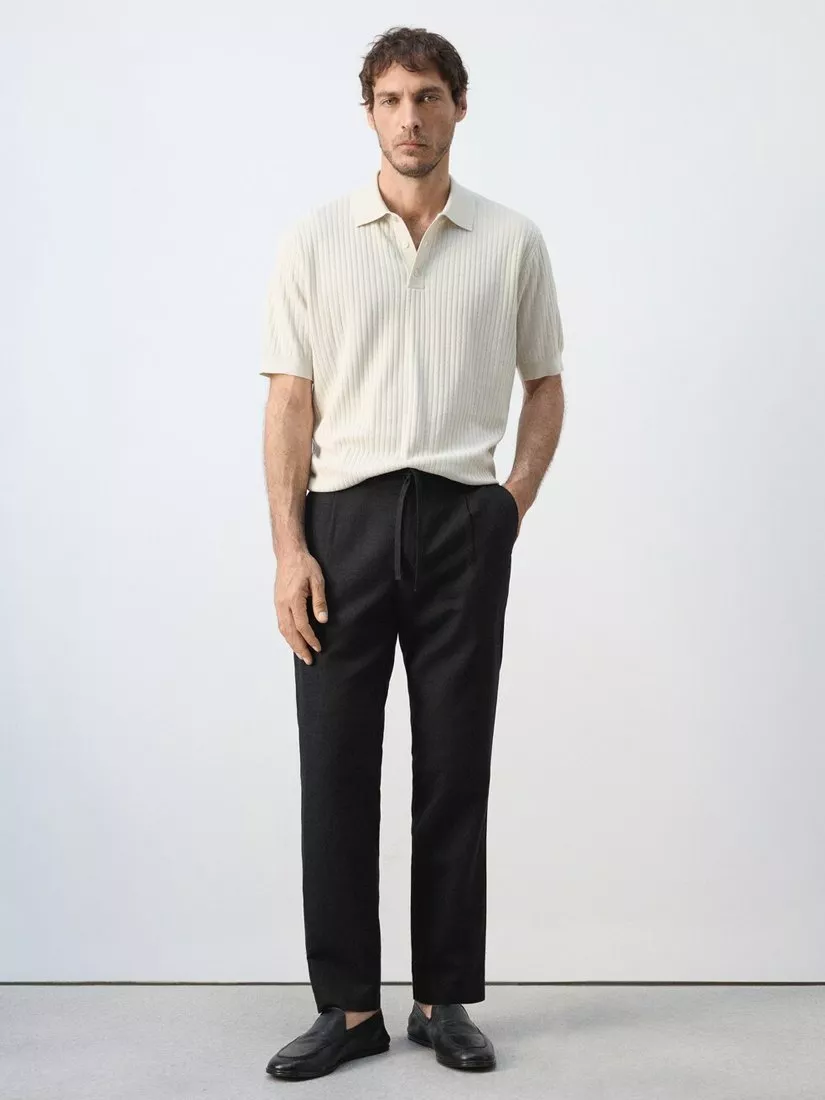 Full-length studio photo of a man wearing a cream ribbed knit polo with black trousers and loafers, minimalist style