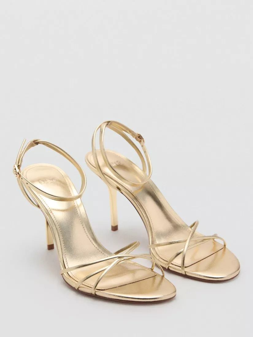 Pair of metallic gold strappy stiletto heel sandals with ankle straps, studio product photo on grey background