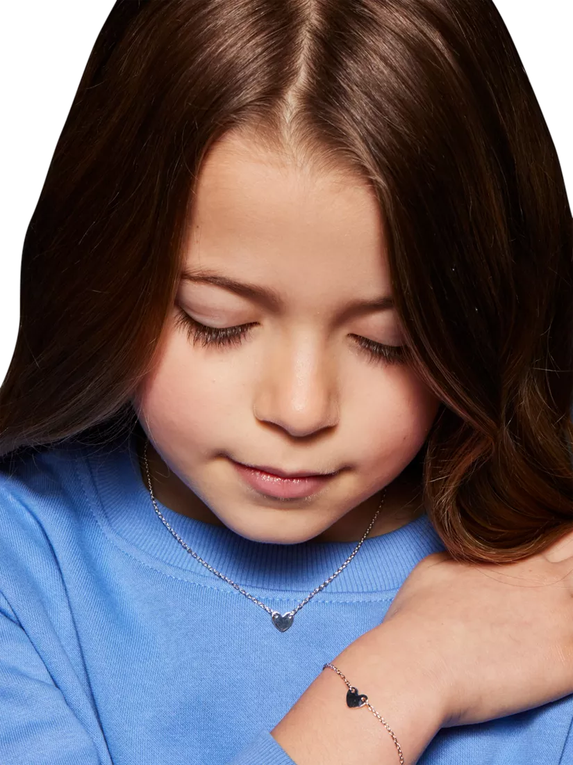 Close-up portrait of young girl in blue jumper wearing heart necklace and bracelet, eyes downcast on white background