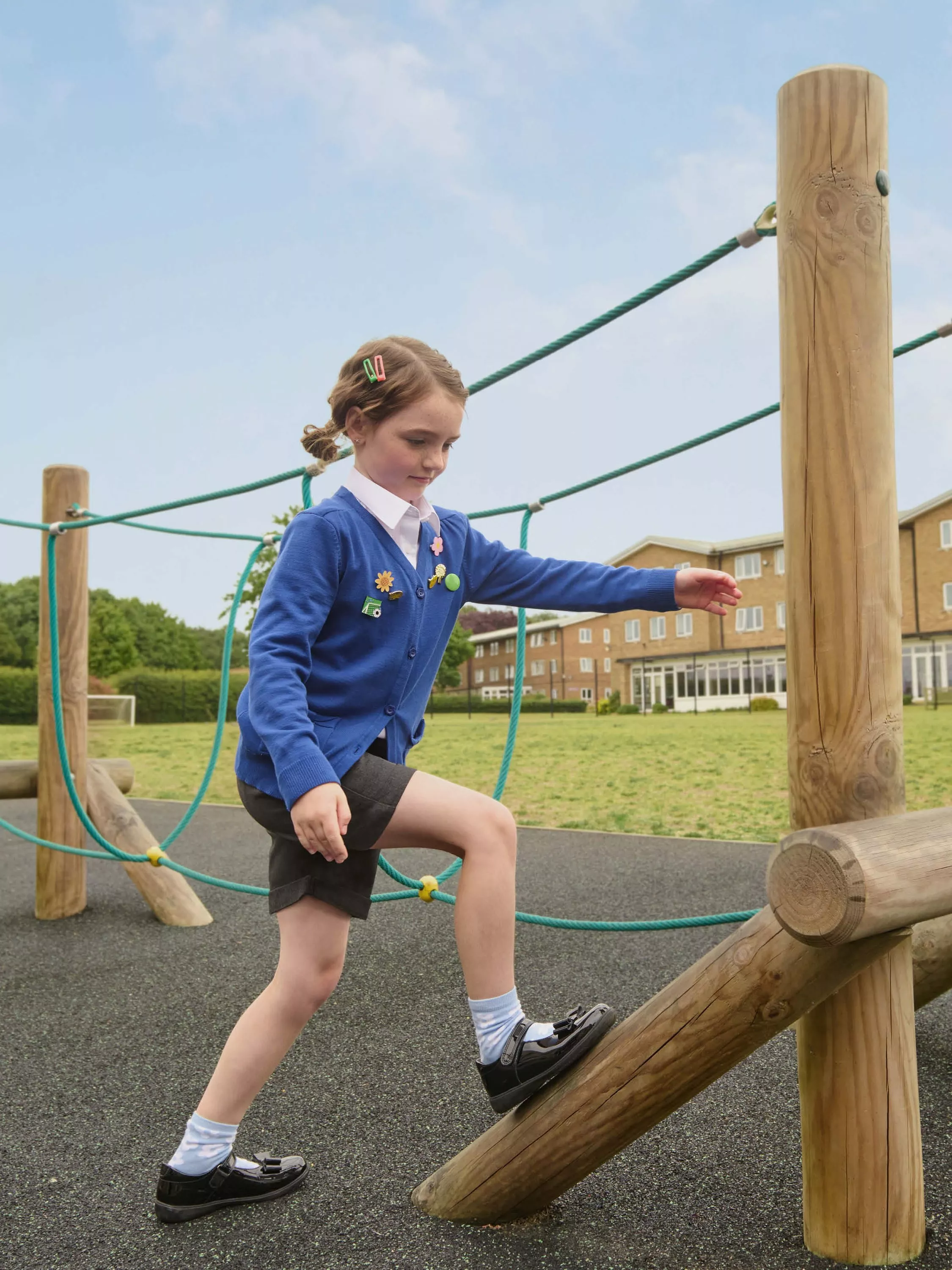 Primary school girl climbing outdoor wooden rope frame on playground, wearing blue uniform cardigan on school field