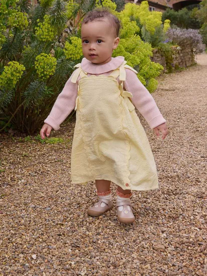 Toddler in yellow pinafore dress and pink top walking on gravel garden path among green shrubs