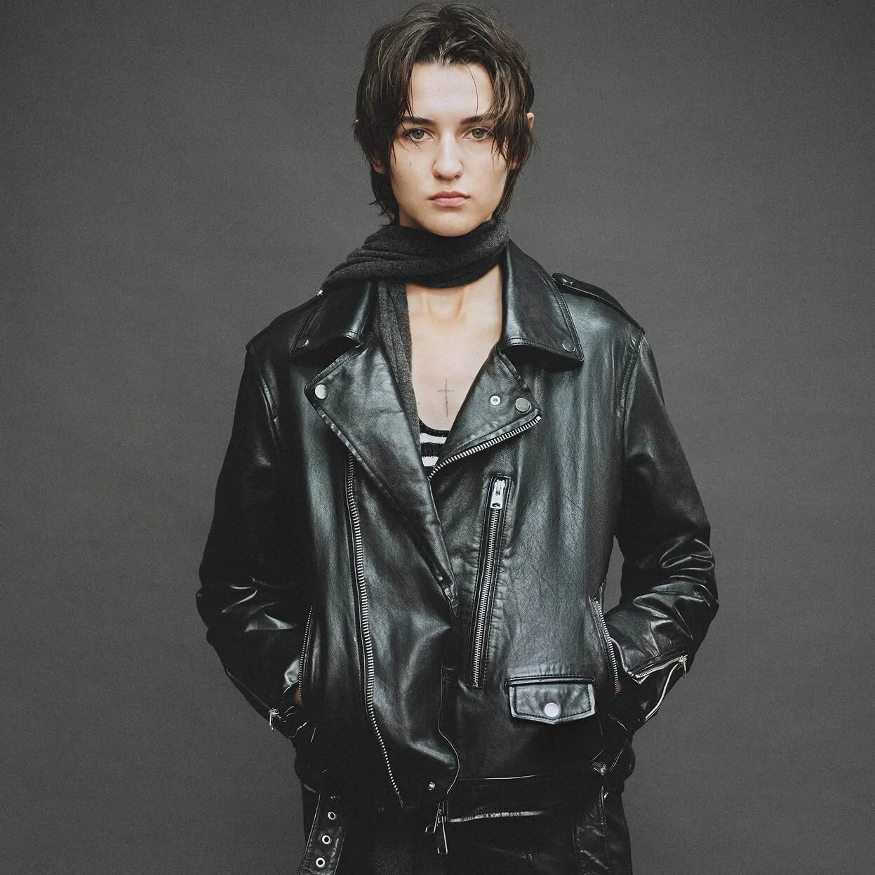 A person with short dark hair wearing a black leather biker jacket and a dark neck scarf against a plain gray studio background.