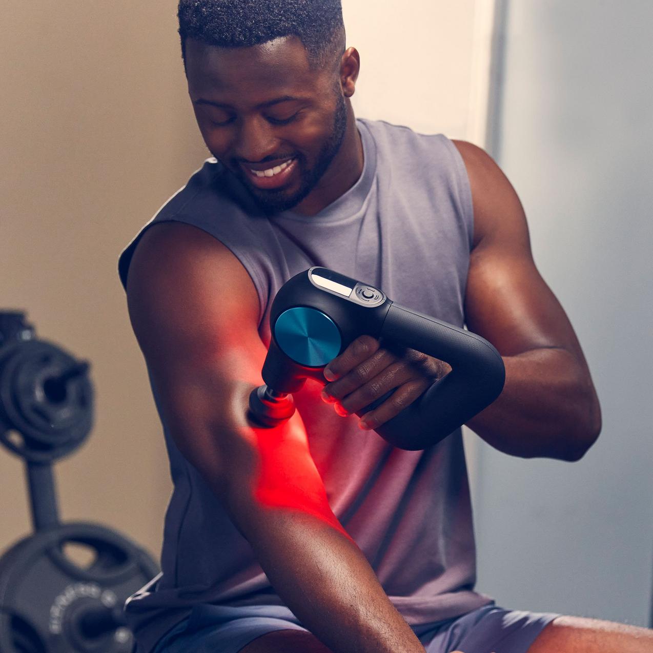 Smiling man using a percussion massage gun with red light therapy on arm for muscle recovery at home gym