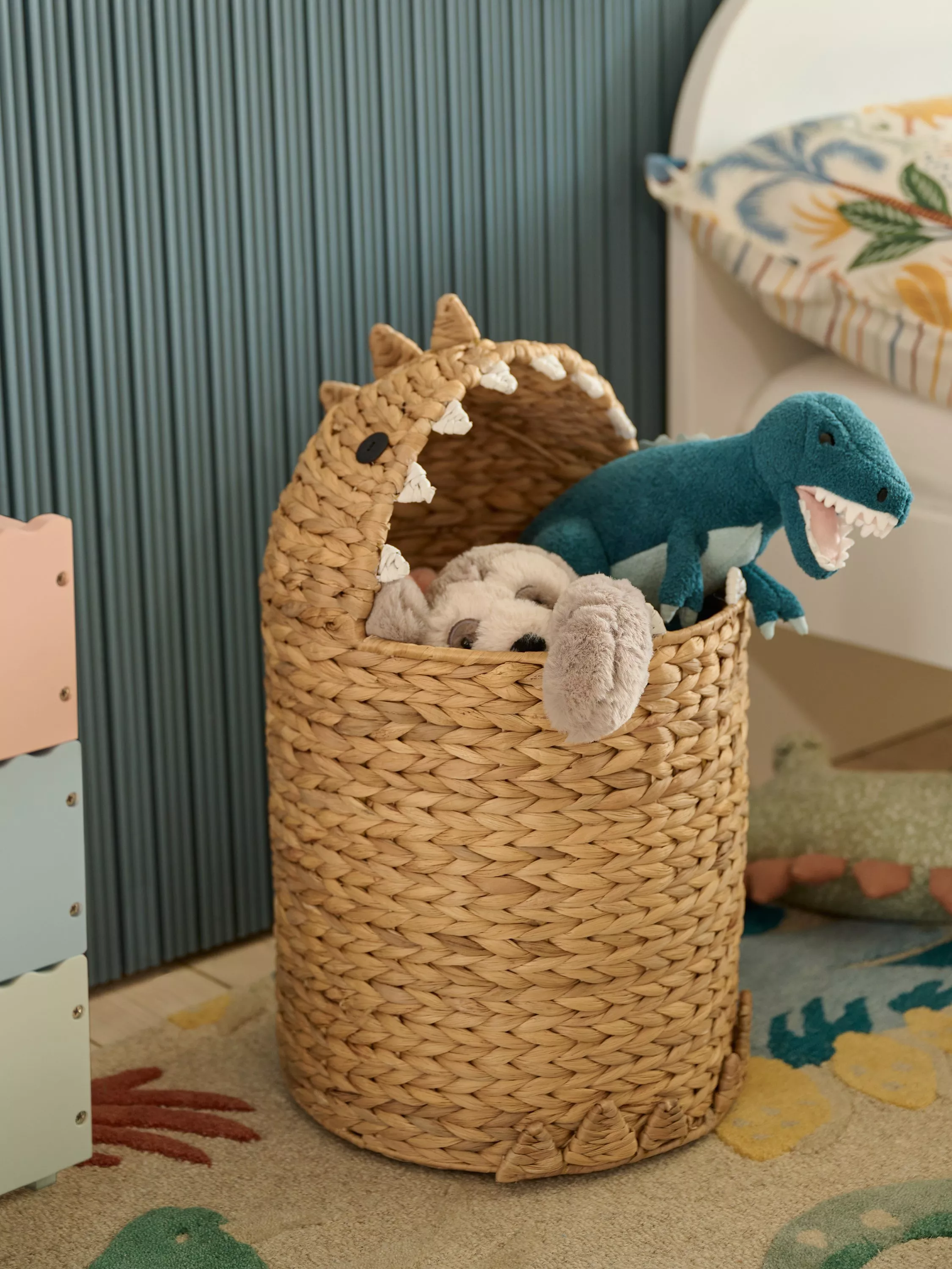 Woven seagrass dinosaur toy storage basket in nursery, filled with soft toys and teal T-rex plush dinosaur
