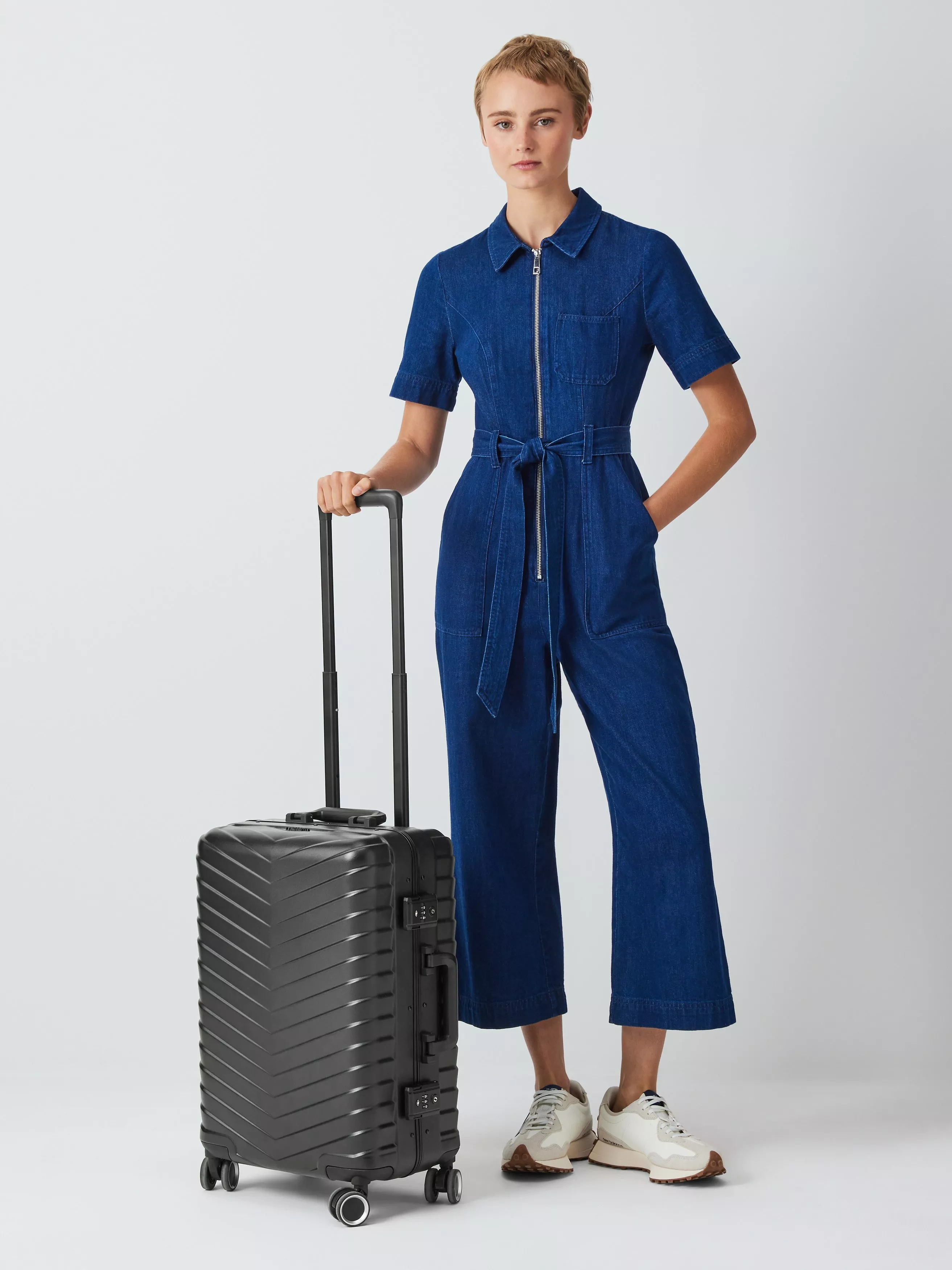 Woman in blue denim jumpsuit standing with black hard-shell spinner suitcase, travel luggage product photo