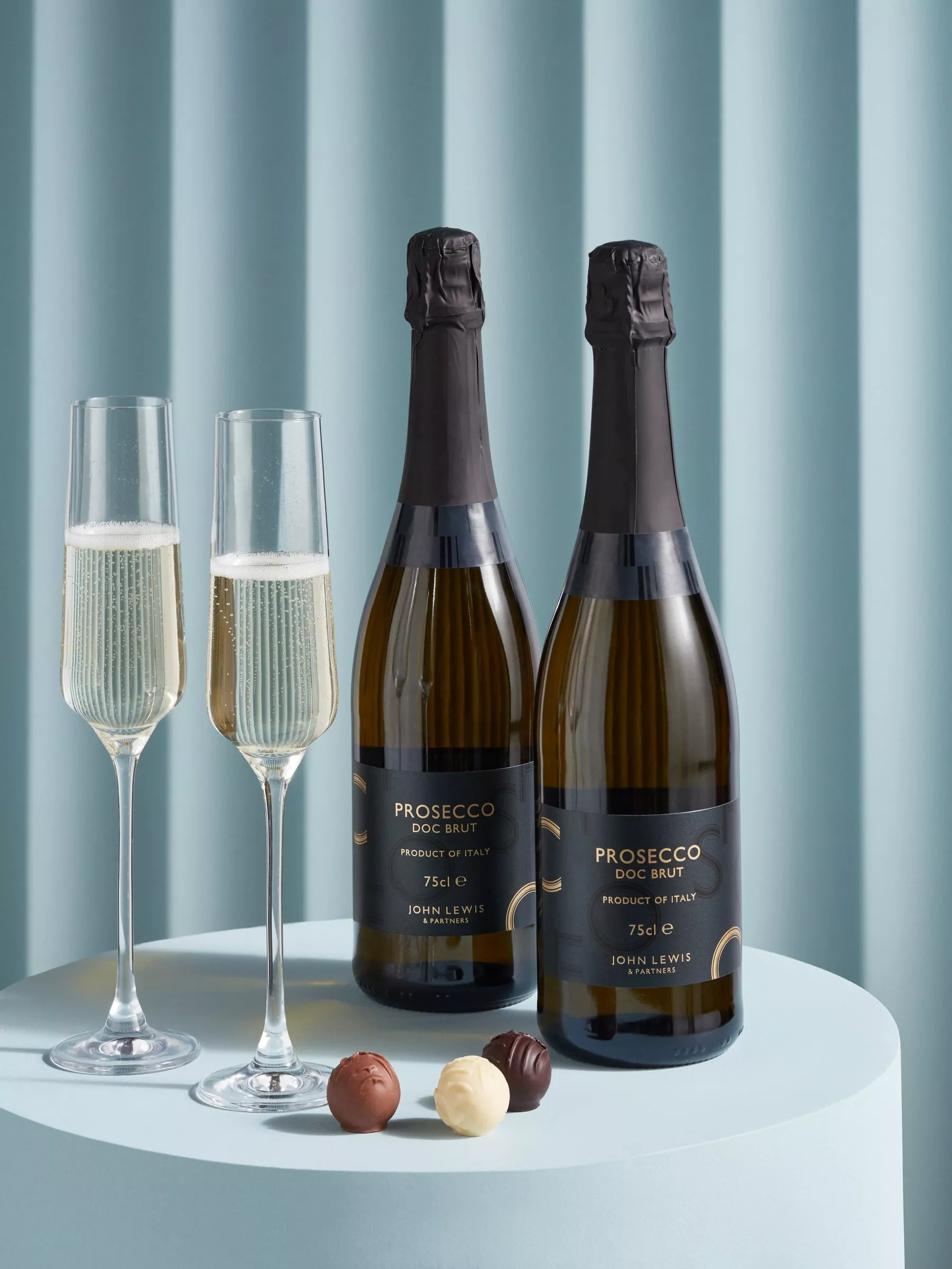 Two bottles of Prosecco DOC Brut with two filled flutes and assorted chocolates on a blue table backdrop