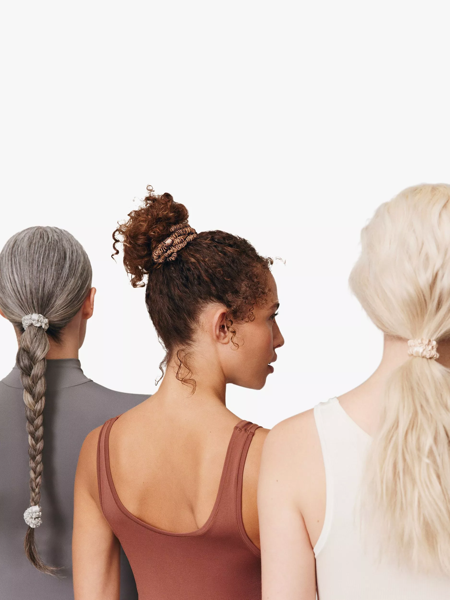Three women from behind showing braided ponytail, curly bun and long blonde ponytail with scrunchies on white background