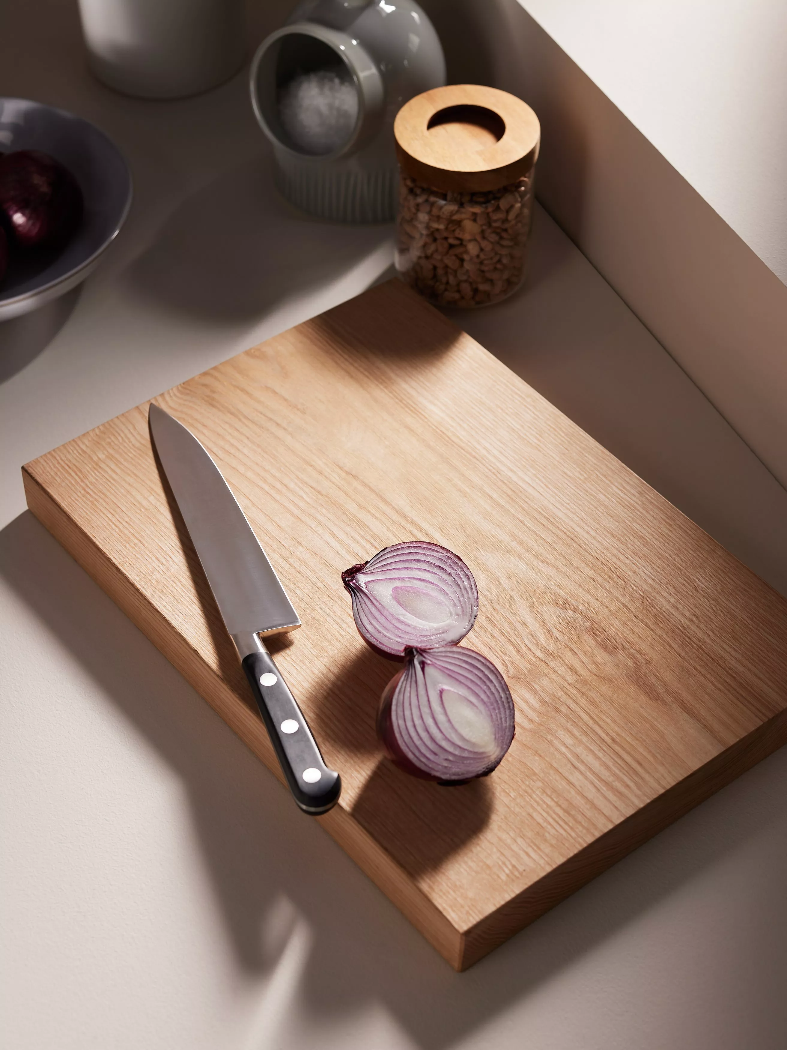 Chef’s knife and halved red onion on wooden chopping board in modern kitchen, with jar and bowl nearby
