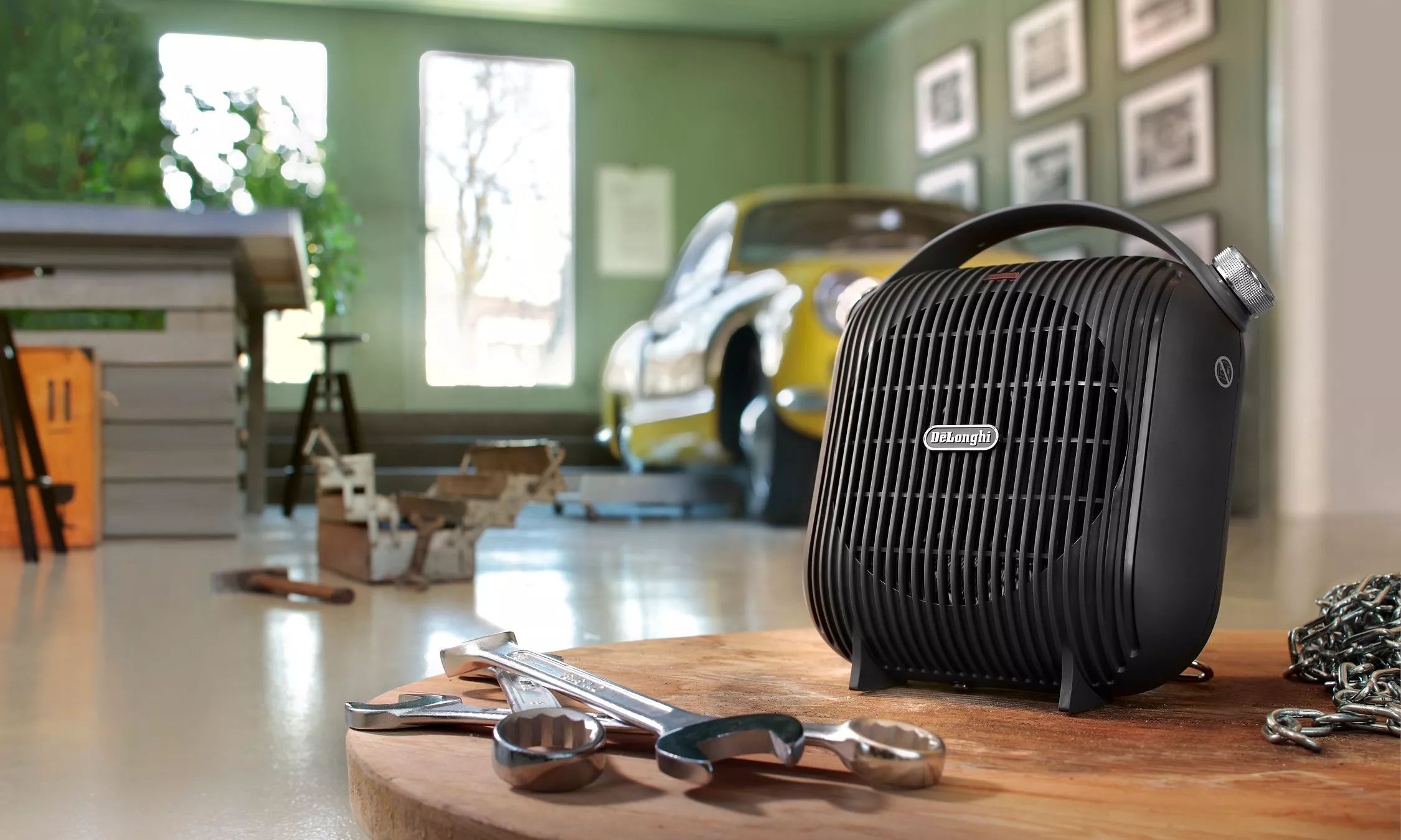 De’Longhi portable fan heater on a workbench with spanners, in a garage workshop with classic car backdrop