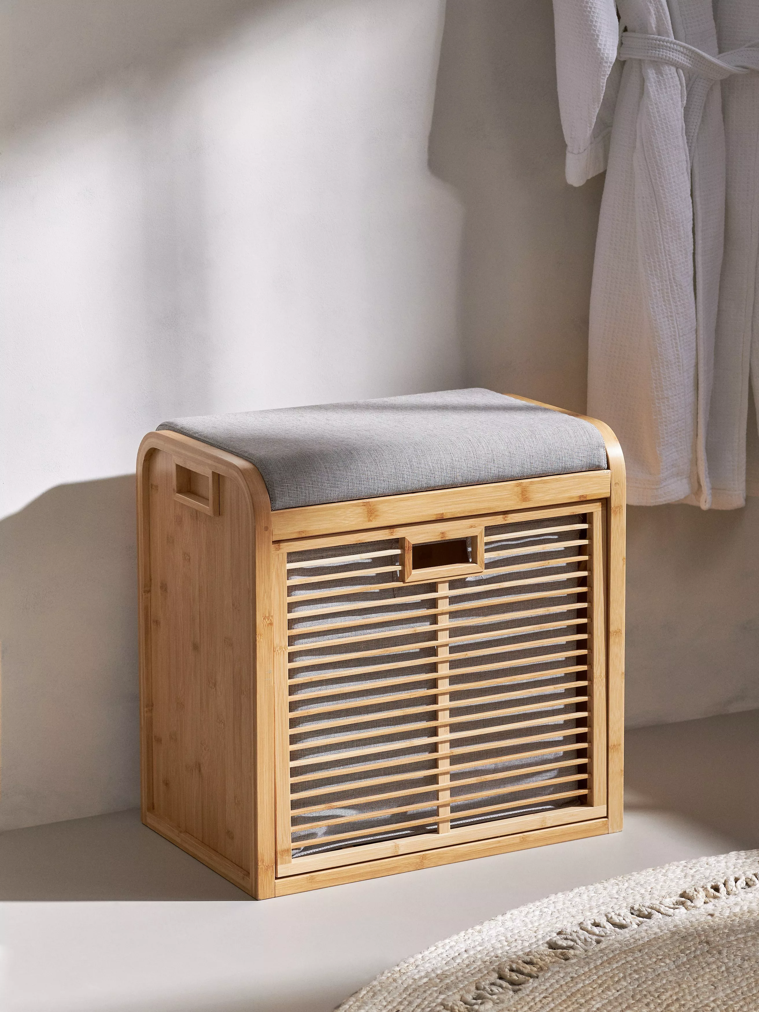 Bamboo bathroom laundry hamper with grey cushioned lid, slatted front and mesh lining beside white bathrobe