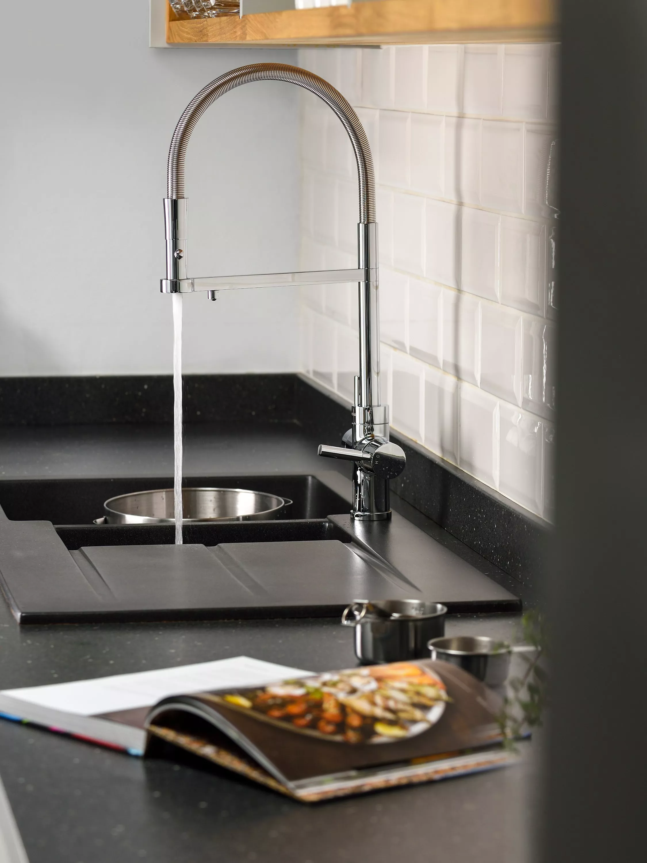 Modern chrome kitchen tap running water over black sink, white tiled splashback and cookbook on worktop