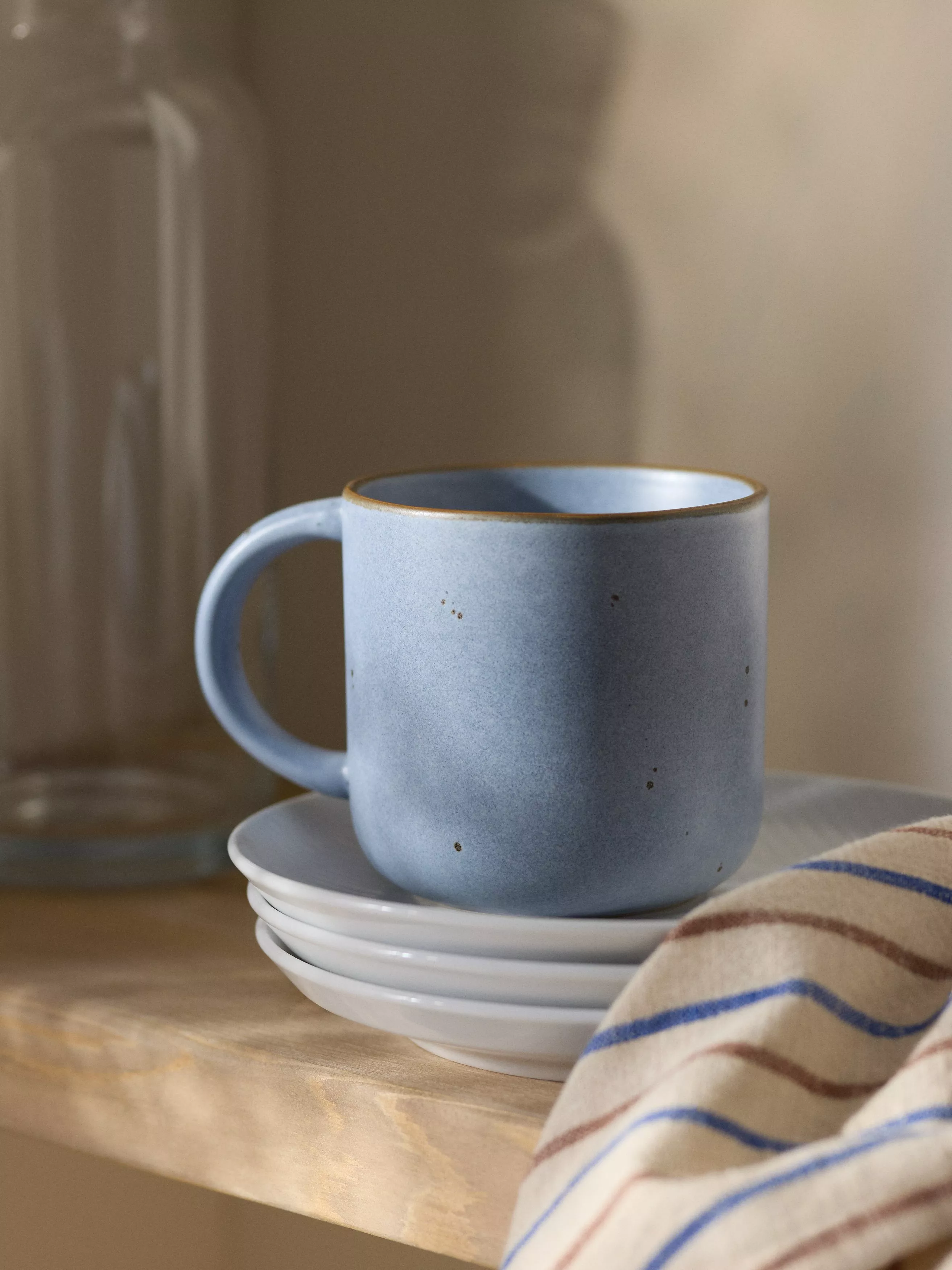 Blue ceramic mug on stacked saucers beside a striped tea towel, cosy kitchen shelf still life close-up