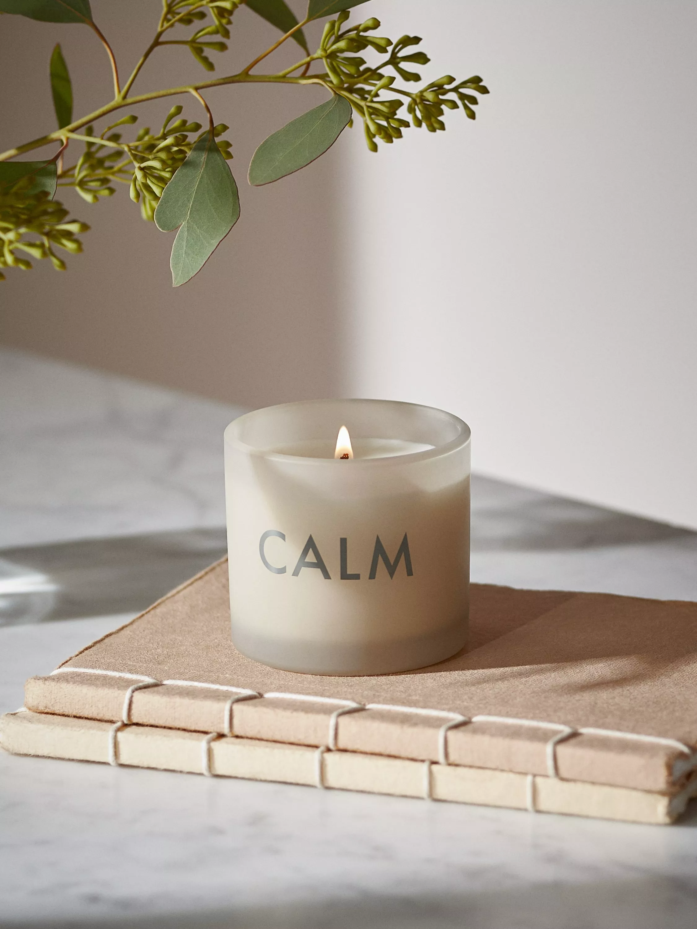 Lit CALM scented candle in frosted glass on a woven tray, with eucalyptus stems in soft natural light