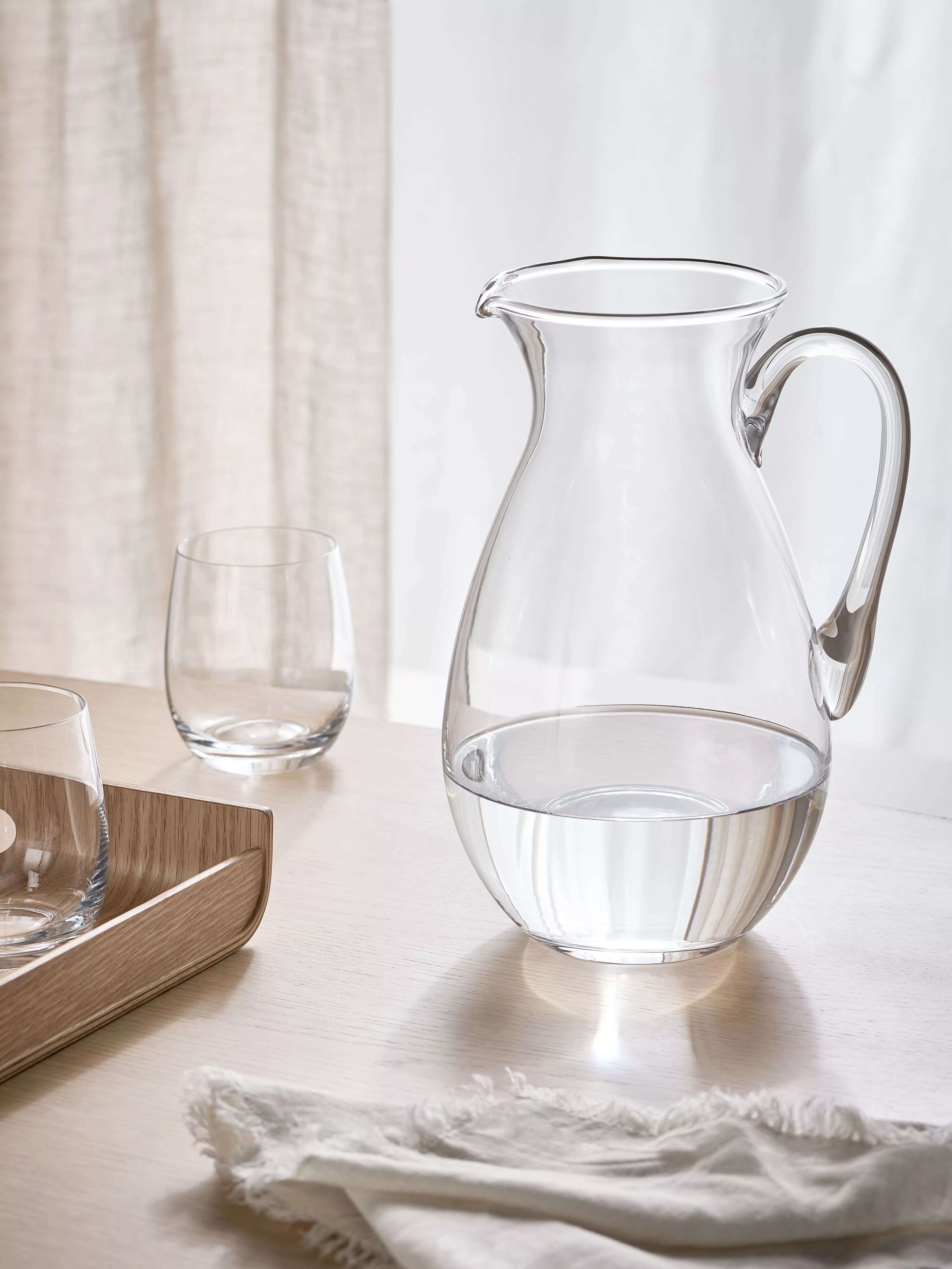 Clear glass water jug and tumblers on light wood table, minimalist dining setting with soft daylight curtains