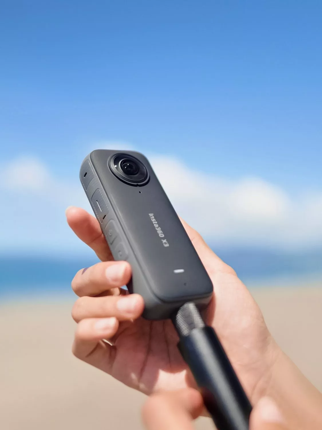 Hand holding Insta360 action camera on selfie stick at beach, blue sky and sea backdrop for travel vlog filming
