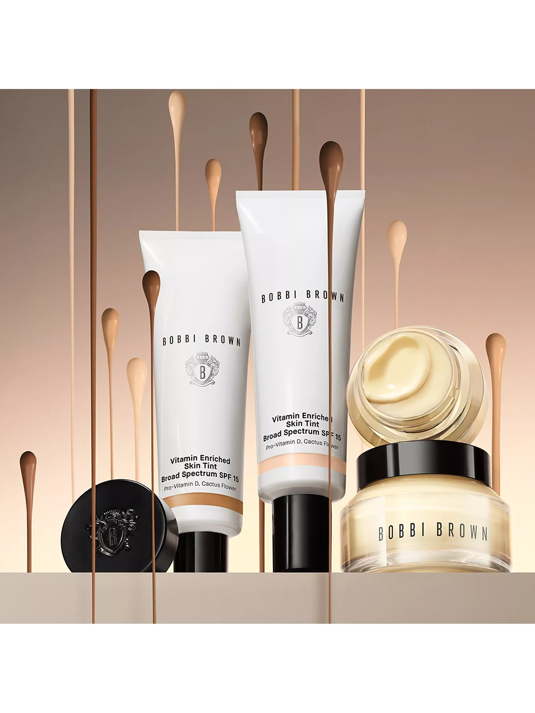 Bobbi Brown Vitamin Enriched Skin Tint SPF 15 tubes and face cream jar with foundation drops on beige backdrop