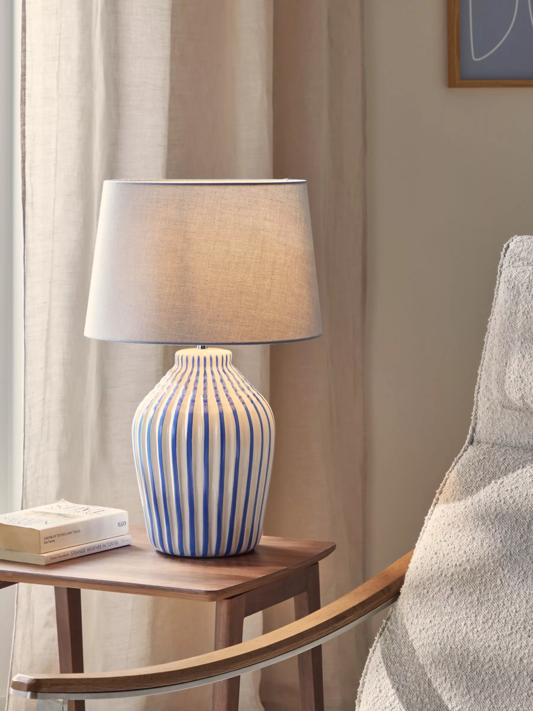 Blue and white striped ceramic table lamp with grey shade on wooden side table beside armchair and curtains
