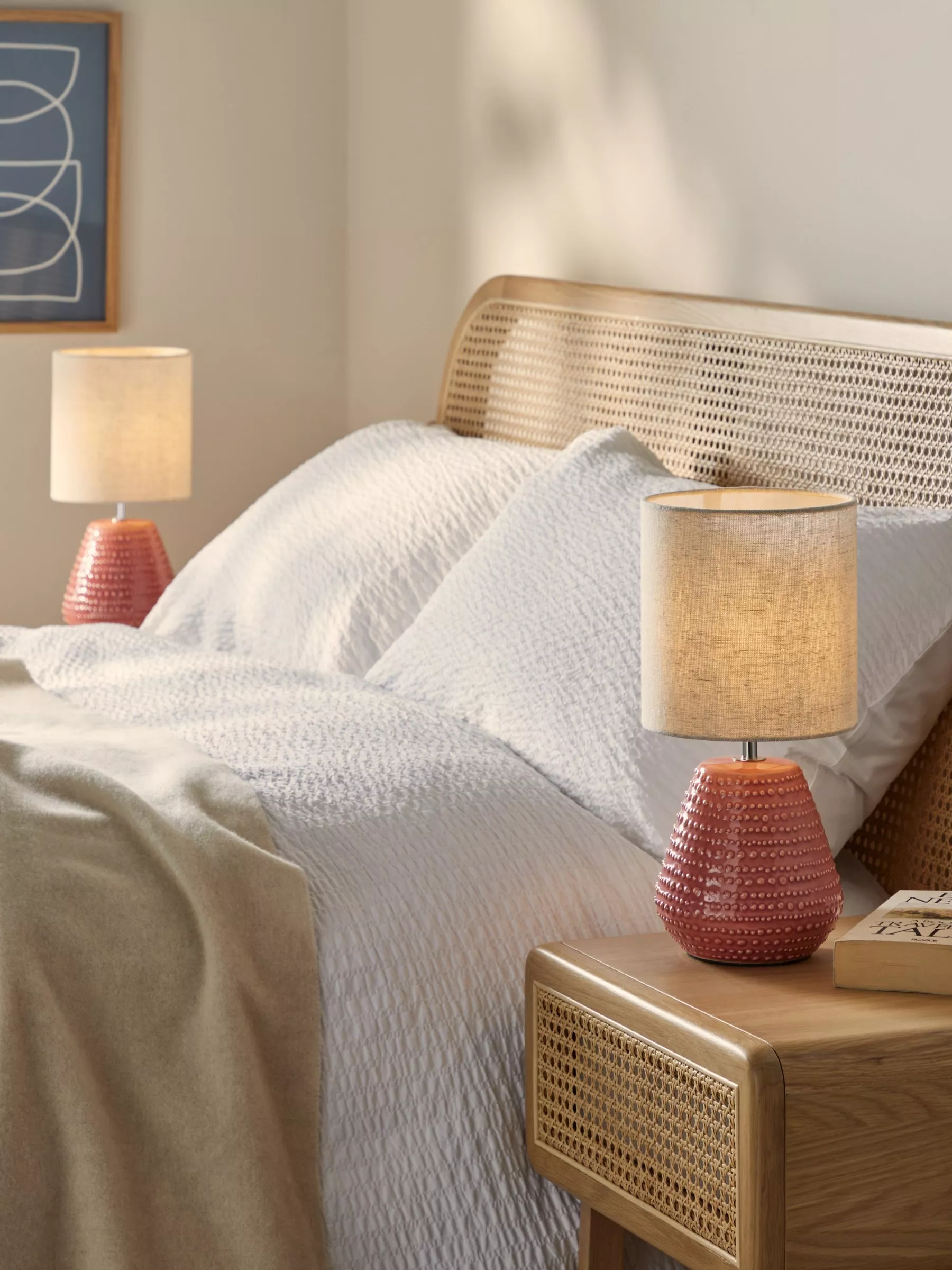 Cosy bedroom with a rattan headboard, white bedding, and pink textured lamps on wooden bedside tables.