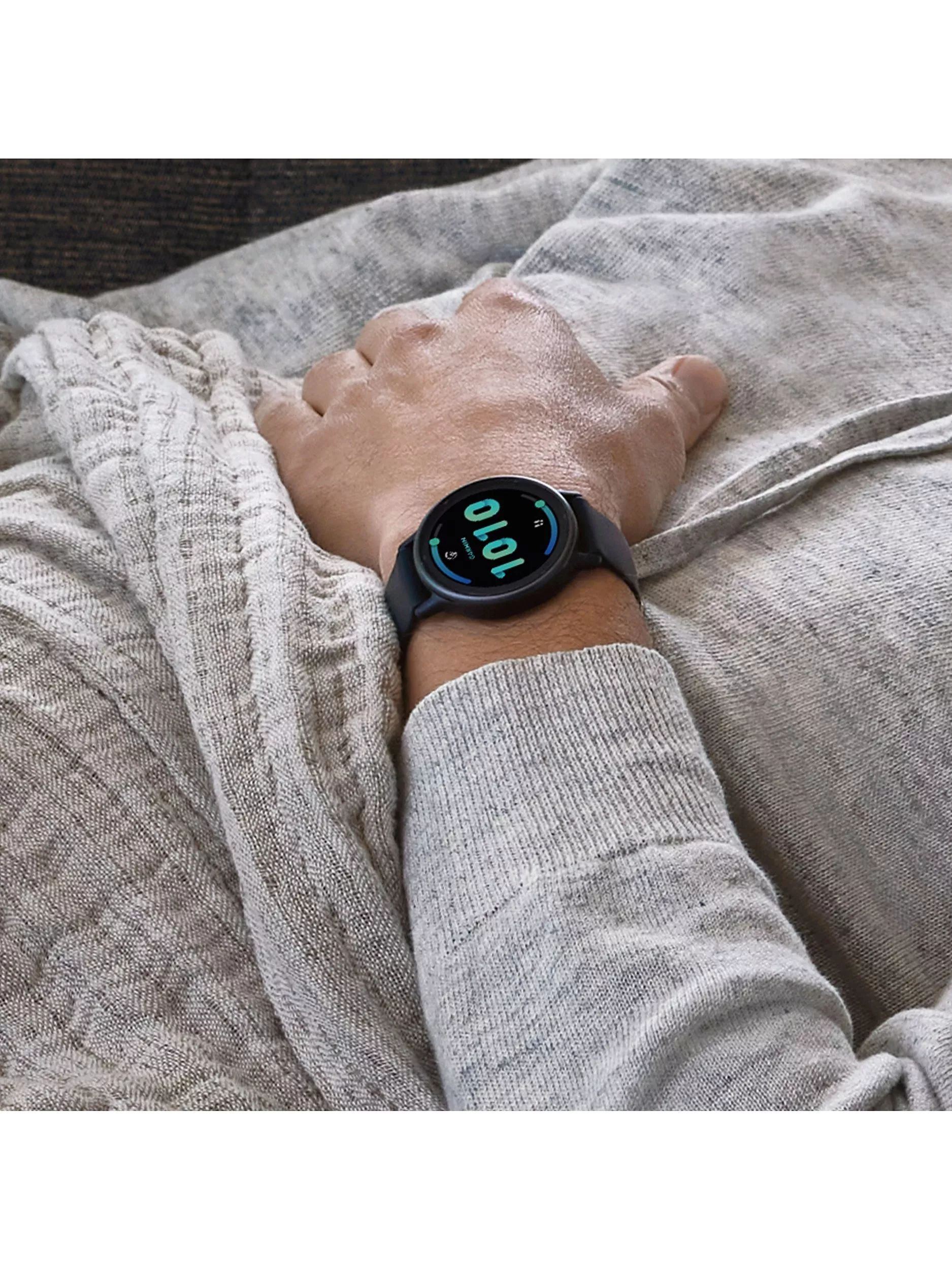 Close-up of wrist wearing a black smartwatch showing 10:10, resting on grey bedding in a bedroom scene