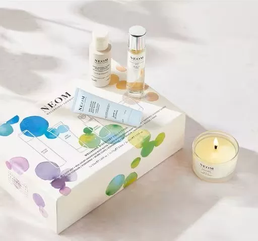 A NEOM wellbeing gift set featuring a collection of skincare bottles, a tube of cream, and a lit scented candle in a clear glass jar.