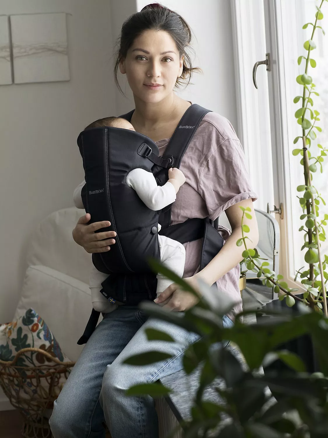 Mother wearing baby in front carrier by window at home, cosy indoor lifestyle photo with natural light