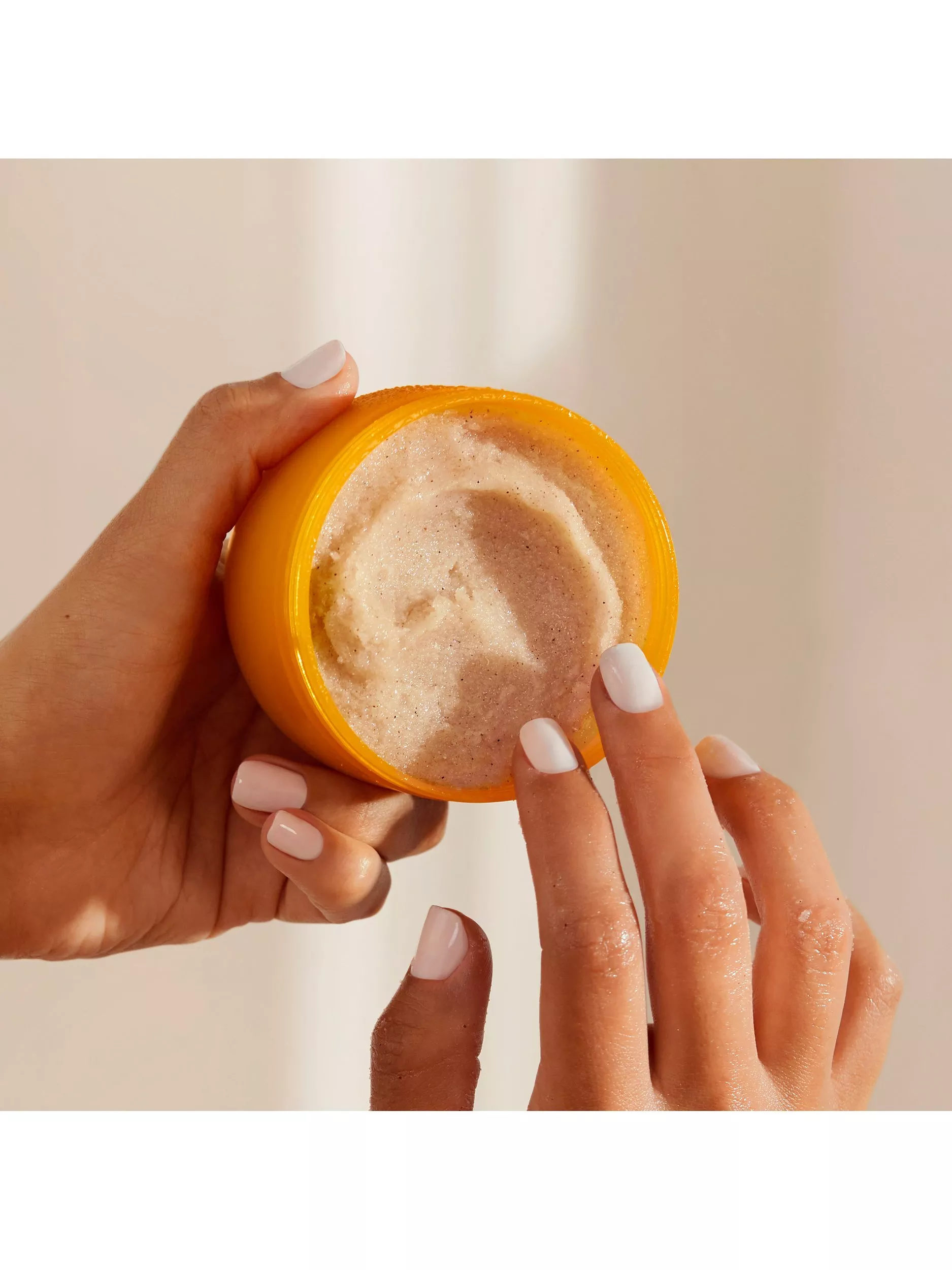 Hands holding open orange jar of exfoliating body scrub, showing creamy texture and manicured nails in soft light