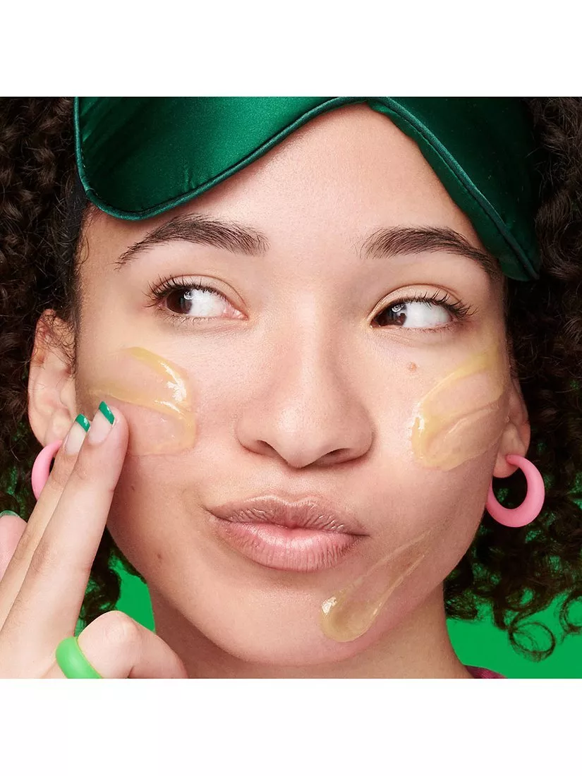 Woman applying face mask with finger, wearing green sleep mask and pink earrings, close-up skincare routine
