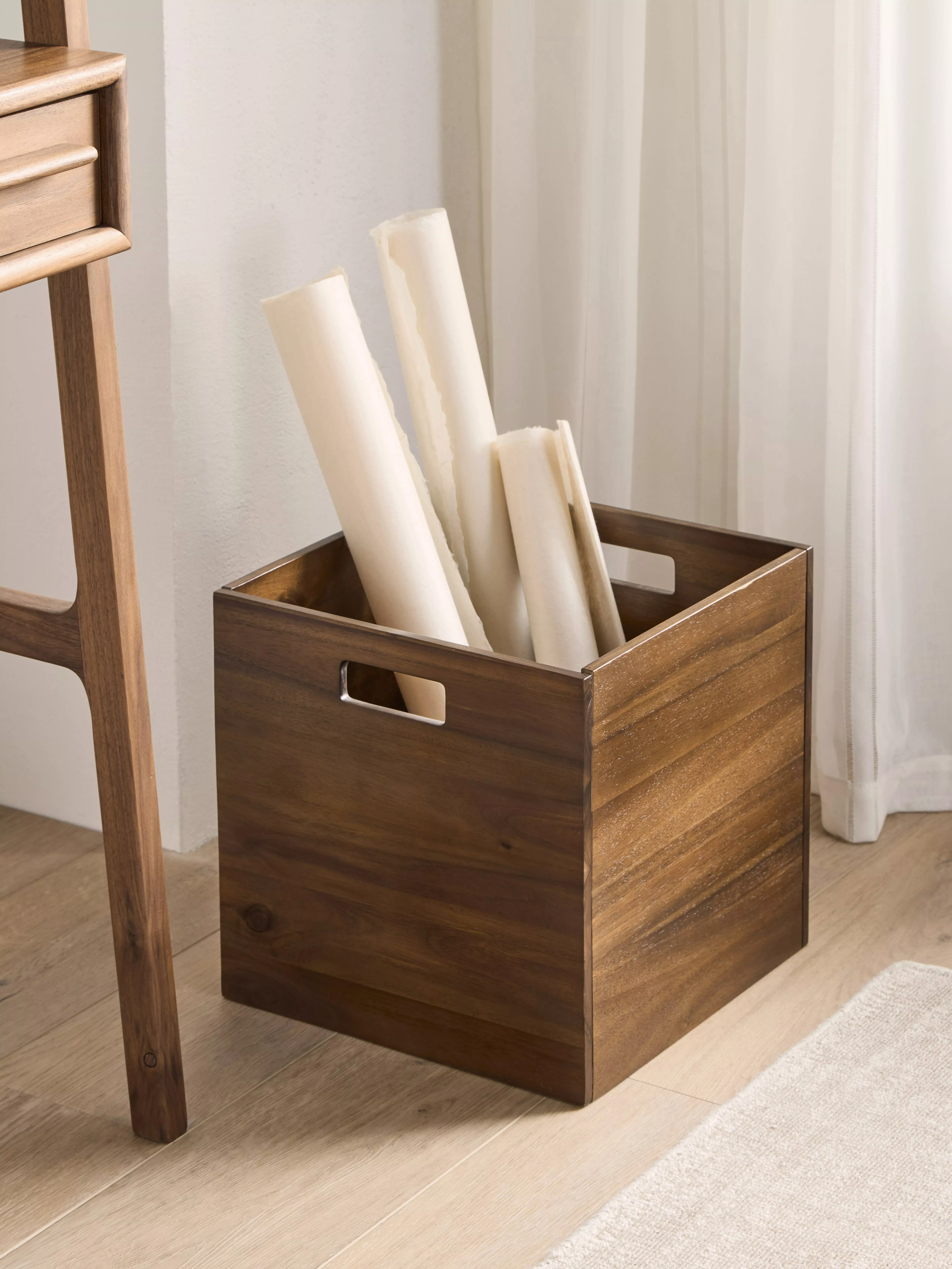 Walnut wooden storage box with handles holding rolled posters, placed by a window in a modern home interior.