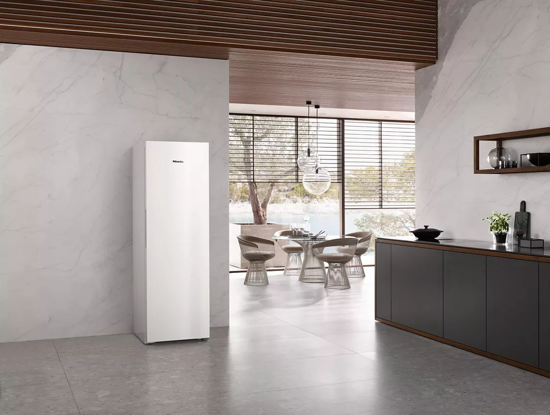 Modern minimalist kitchen with tall white refrigerator, marble walls, dark cabinets, and dining area by large window.