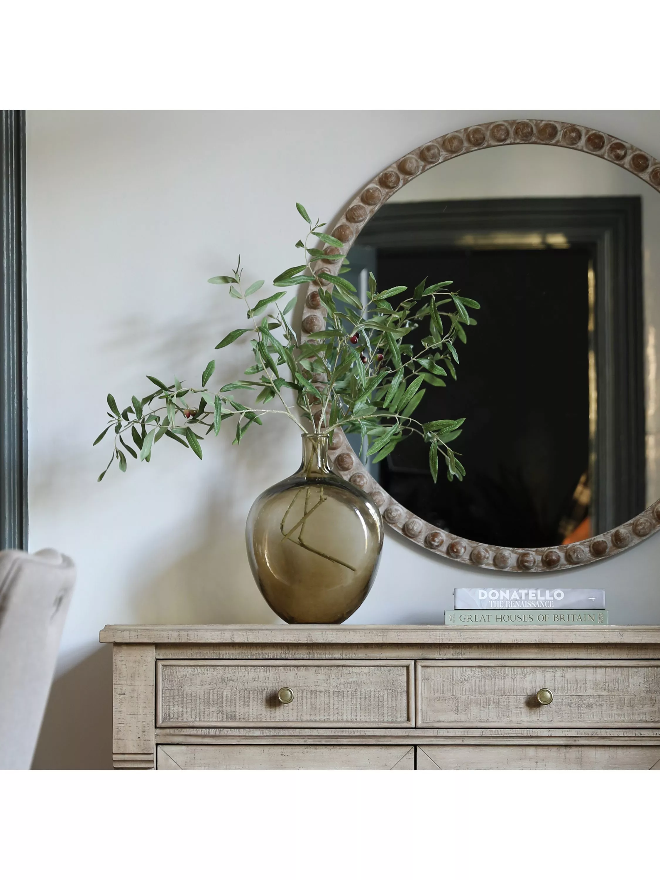 Rustic wooden sideboard with round framed mirror, glass vase of olive branches, and stacked books in cosy home decor