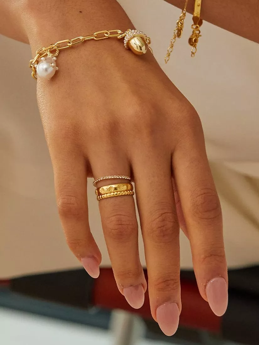 Hand with 3 gold rings stack on one finger and a gold bracelet, with pink nail varnish
