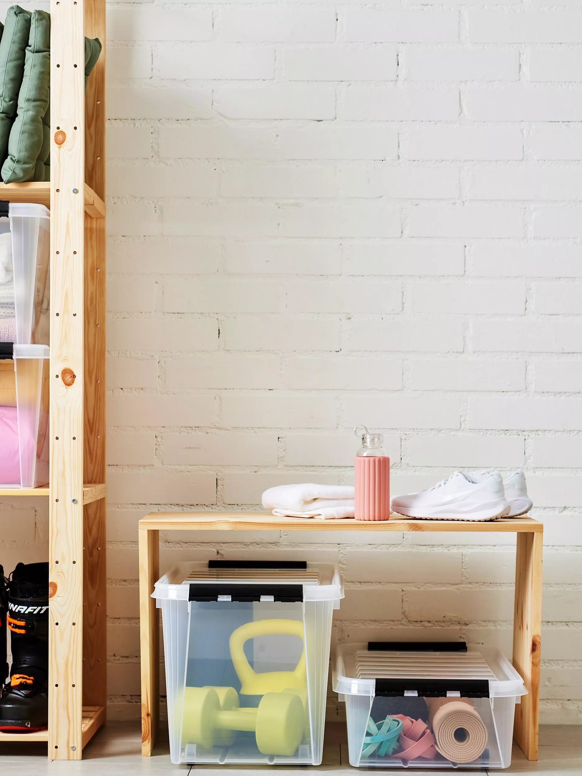 Home gym storage corner with wooden shelf, trainers, towel, drink bottle, and clear boxes of weights and mats