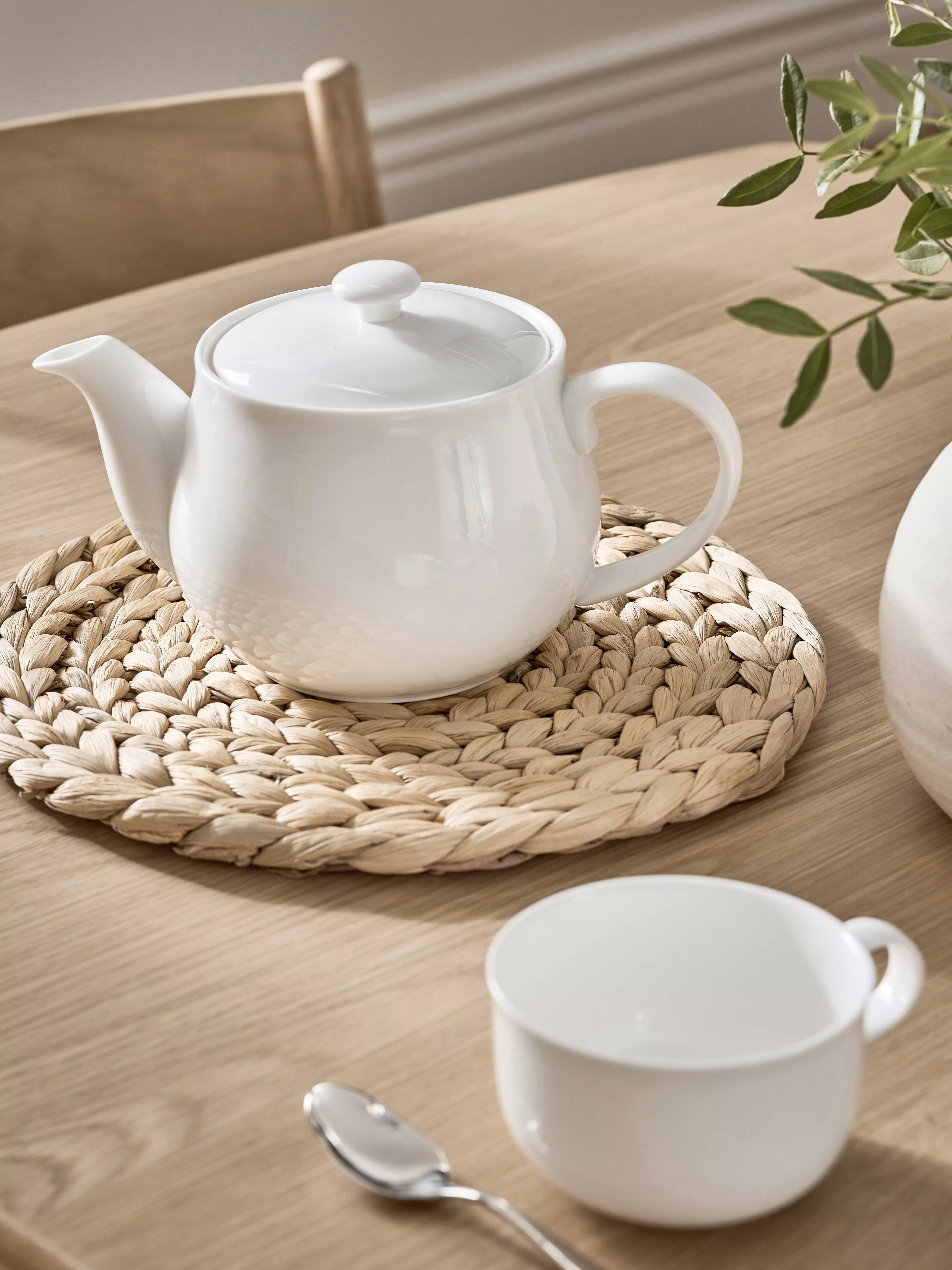 White ceramic teapot on woven placemat with teacup on wooden table, minimalist kitchen dining setting